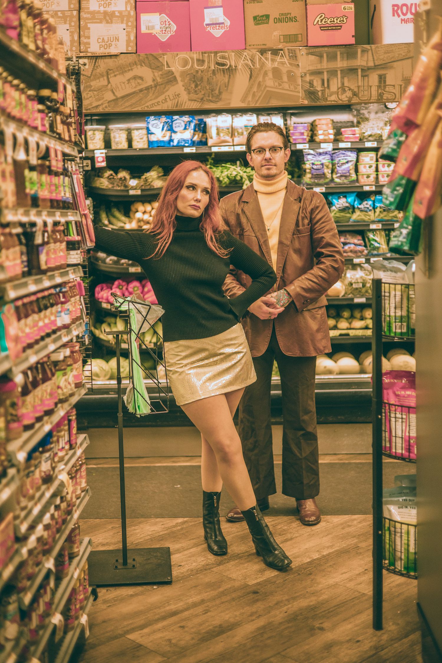 A woman in a gold skirt and man in brown blazer pose in a colorful grocery store aisle.