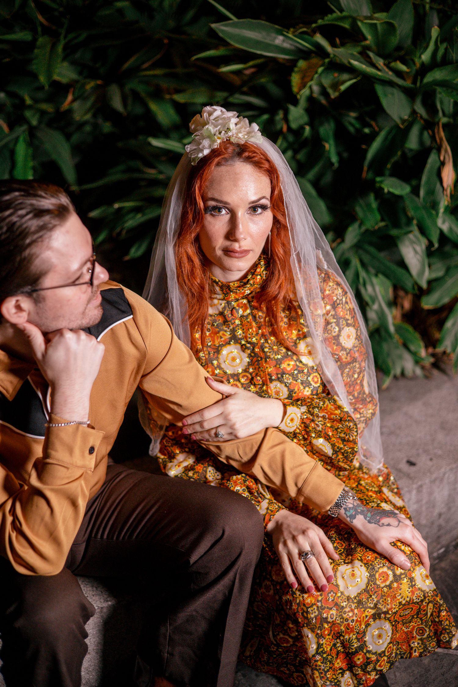 A redheaded bride in a floral dress and veil sits with a man in a mustard jacket among lush green foliage.