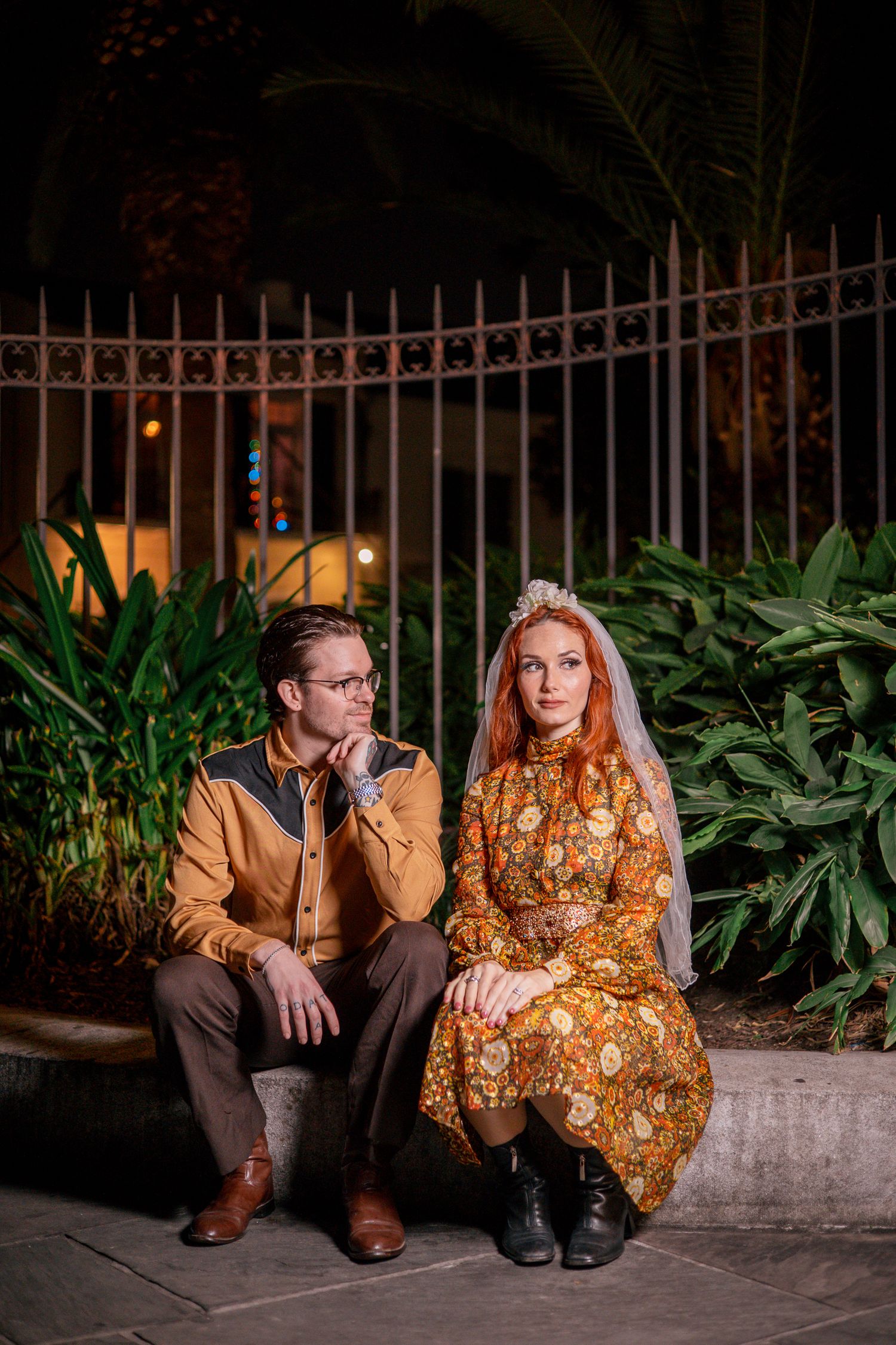 Couple in vintage-style outfits sitting outdoors at night, bride in floral dress with veil and groom in brown jacket.