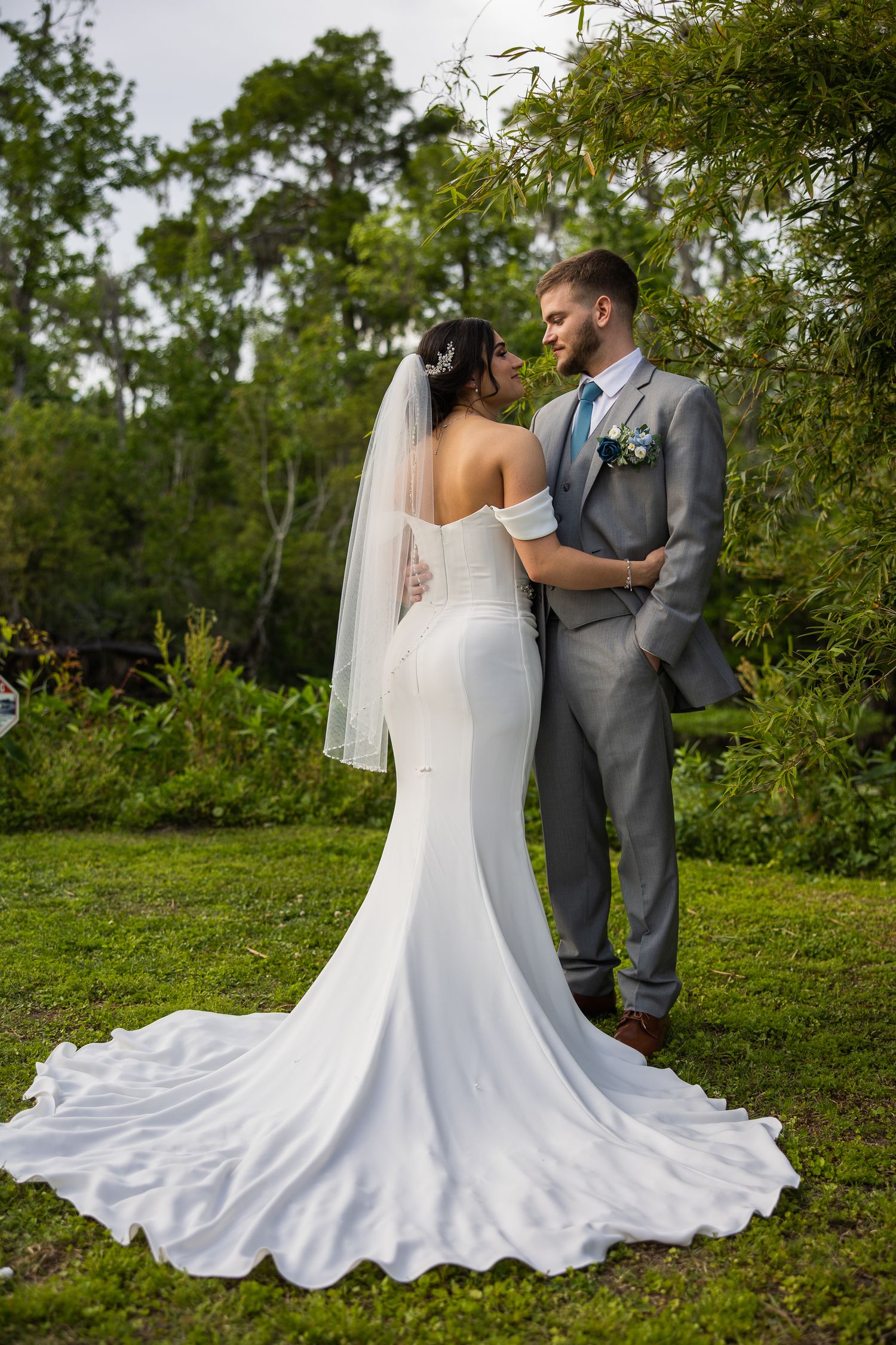 Bride in fitted mermaid gown with veil embraces groom in gray suit outdoors in garden.