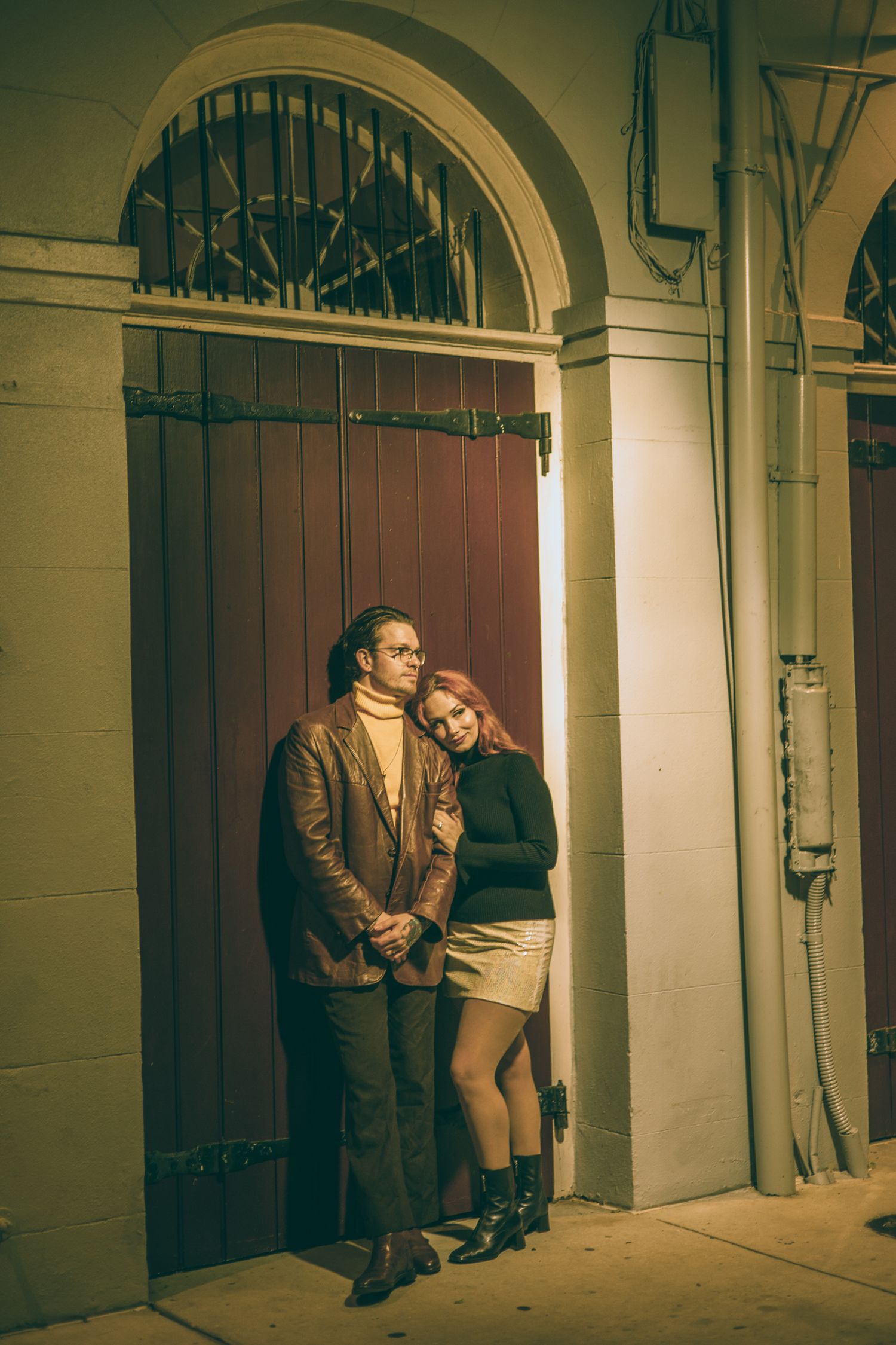 Couple posing at night by a vintage wooden door with arched window, woman in black top leaning on man in leather jacket.
