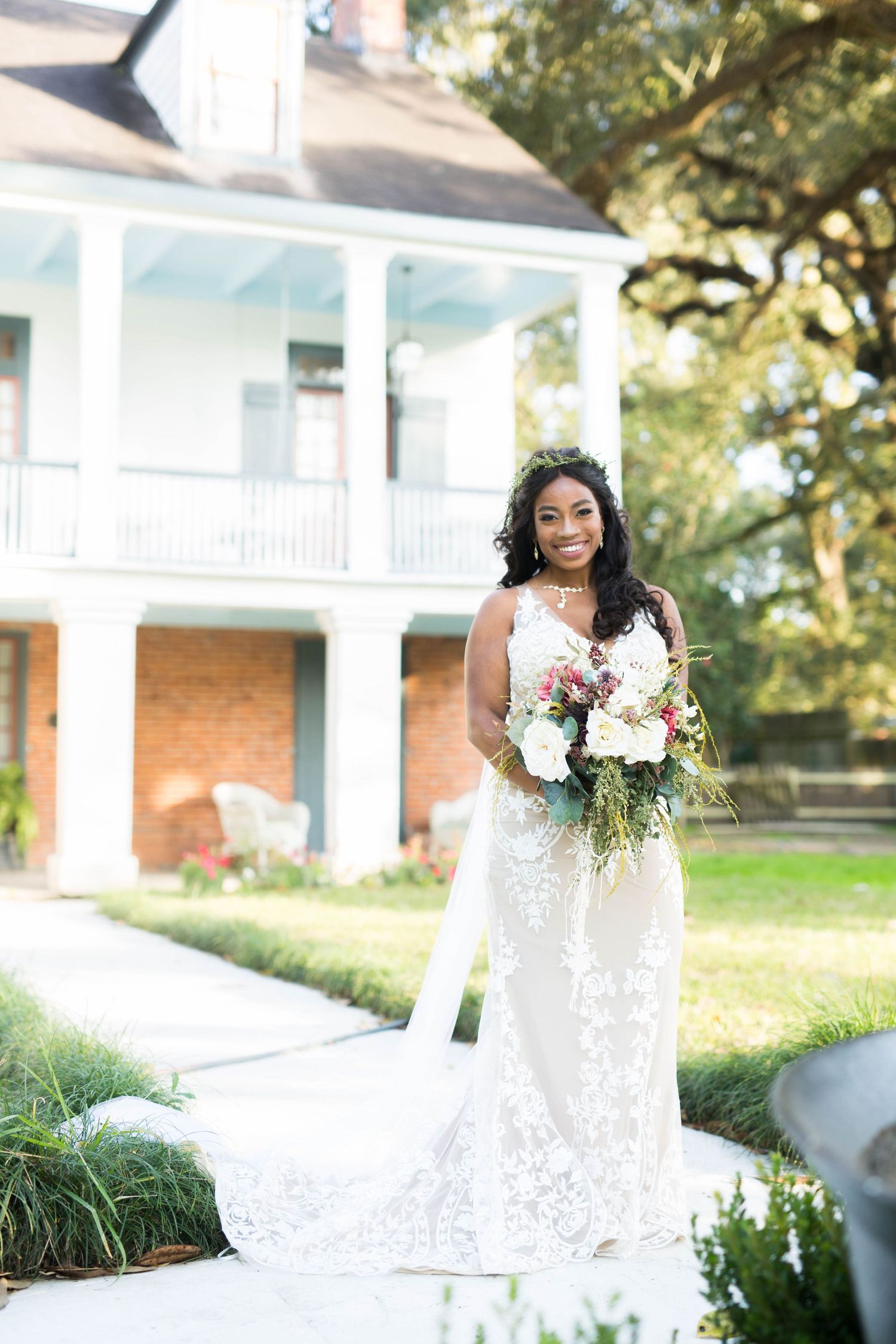 Smiling bride in lace wedding gown holds bouquet outside a white Southern plantation home.
