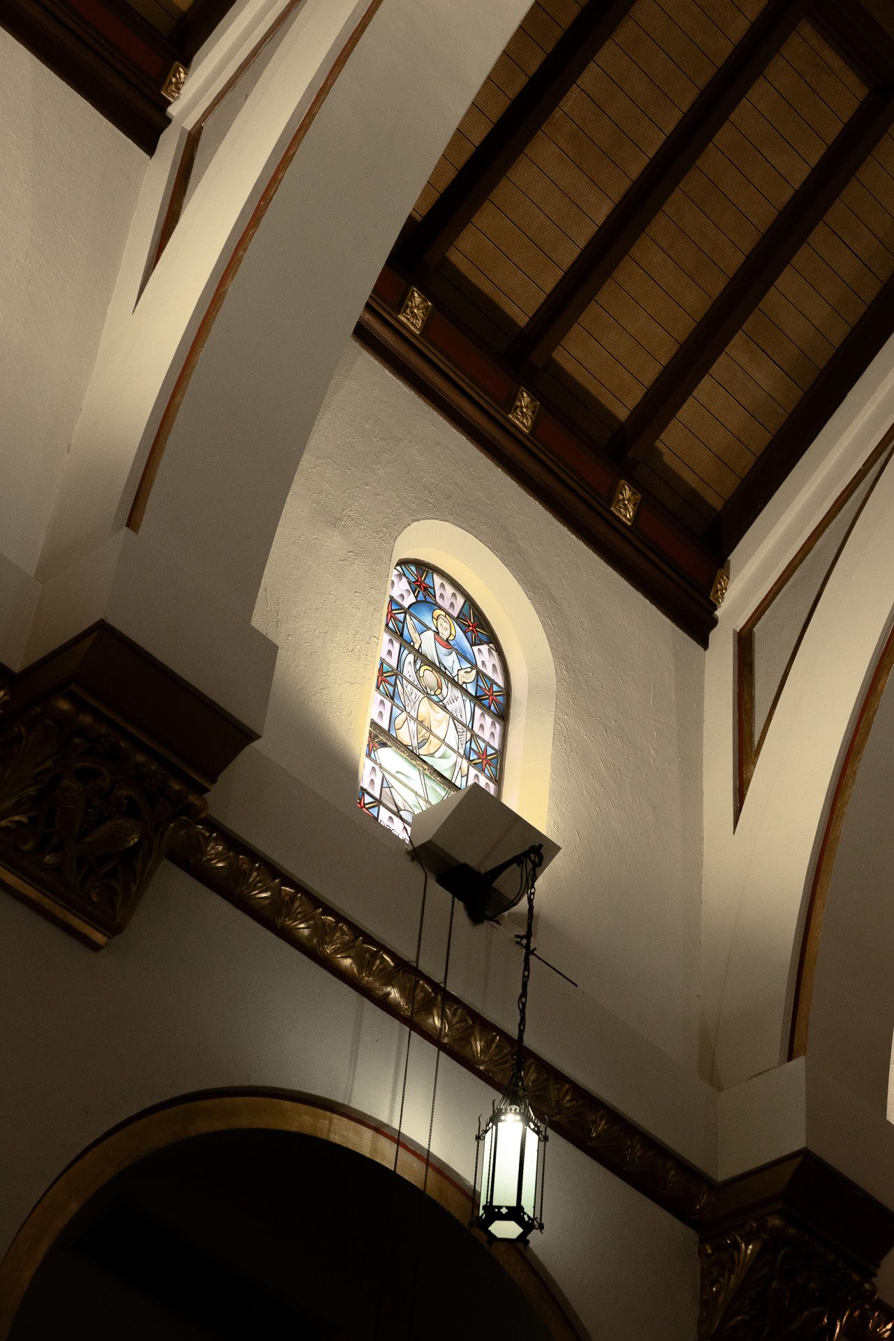 Interior church ceiling with wooden beams, arched stained glass window, and hanging lantern in warm light.