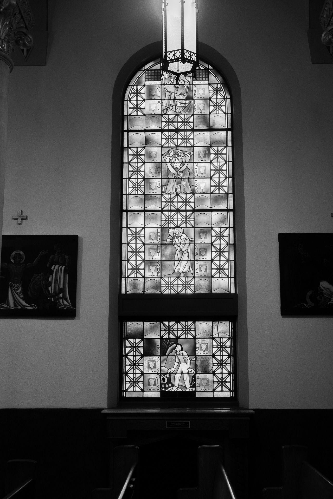 Black and white photo of a tall arched stained glass window in a church interior with dark wood pews and religious artwork.