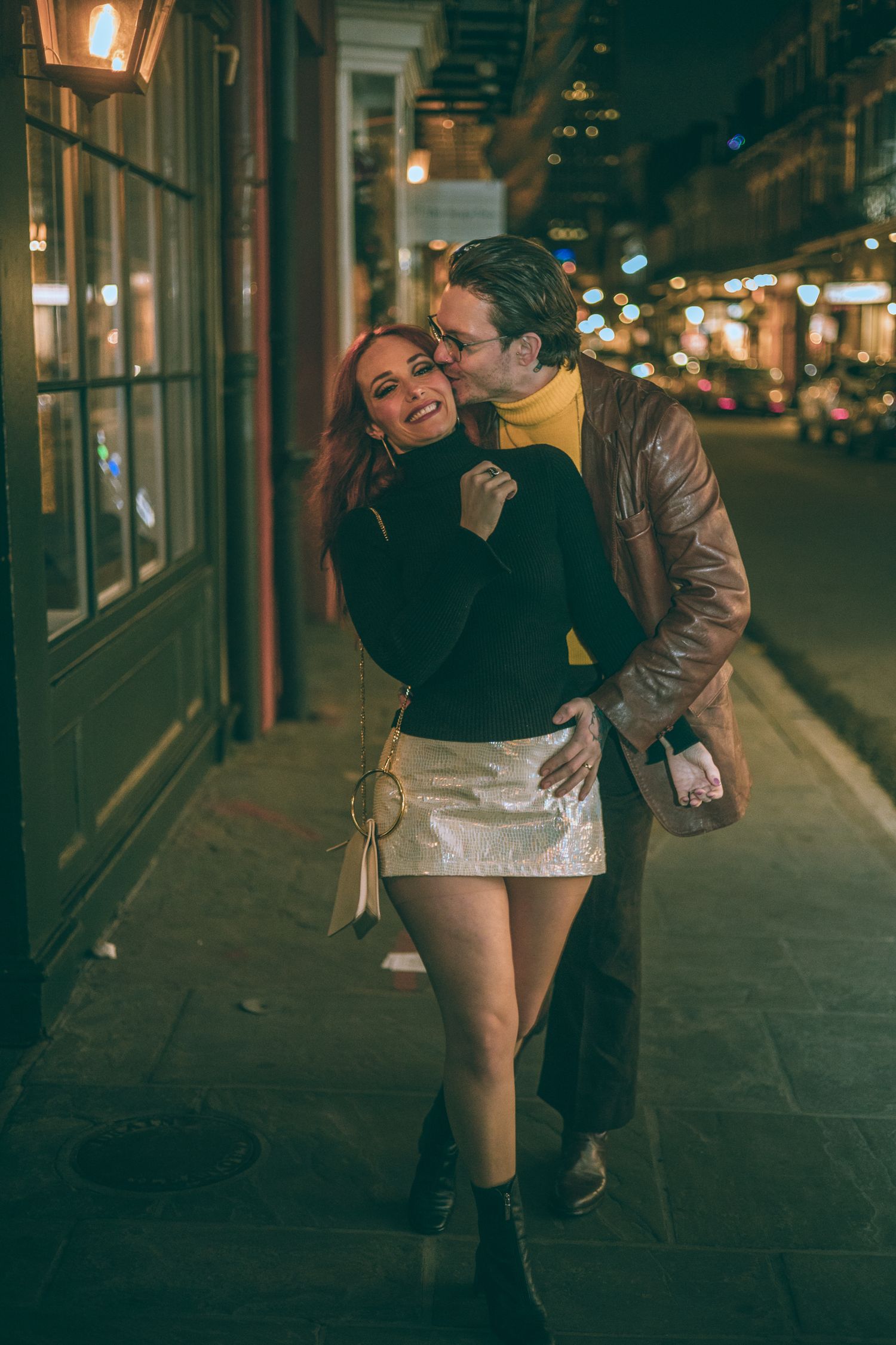 Couple sharing a romantic moment on a city street at night, woman in black sweater and white skirt smiling.