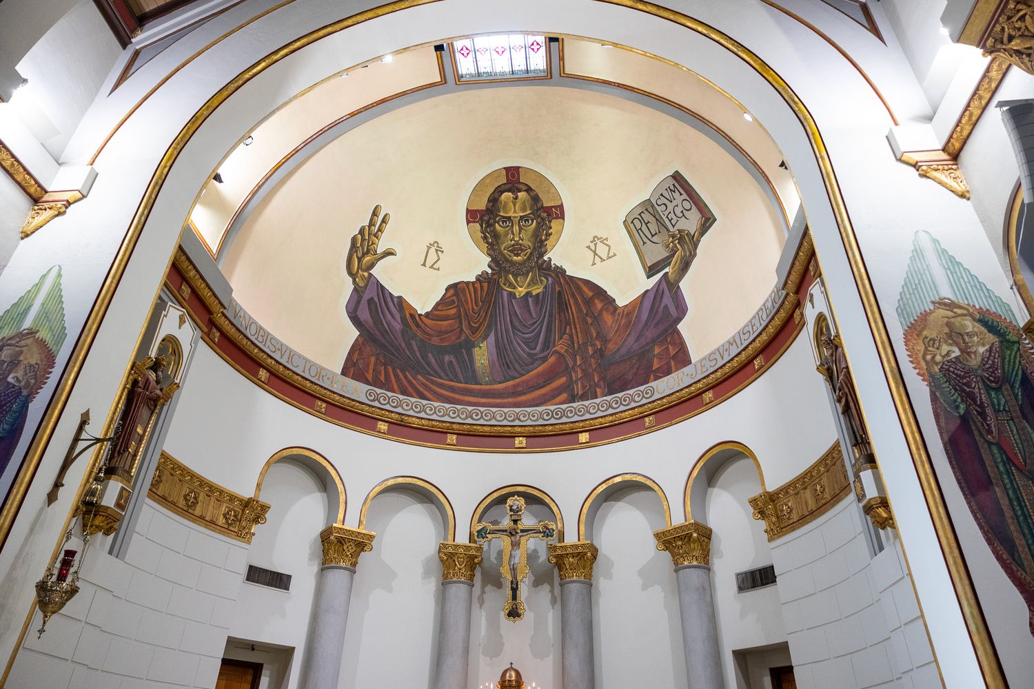 Interior church apse with mosaic of Christ Pantocrator, gold accents, arched alcoves, and a crucifix below.
