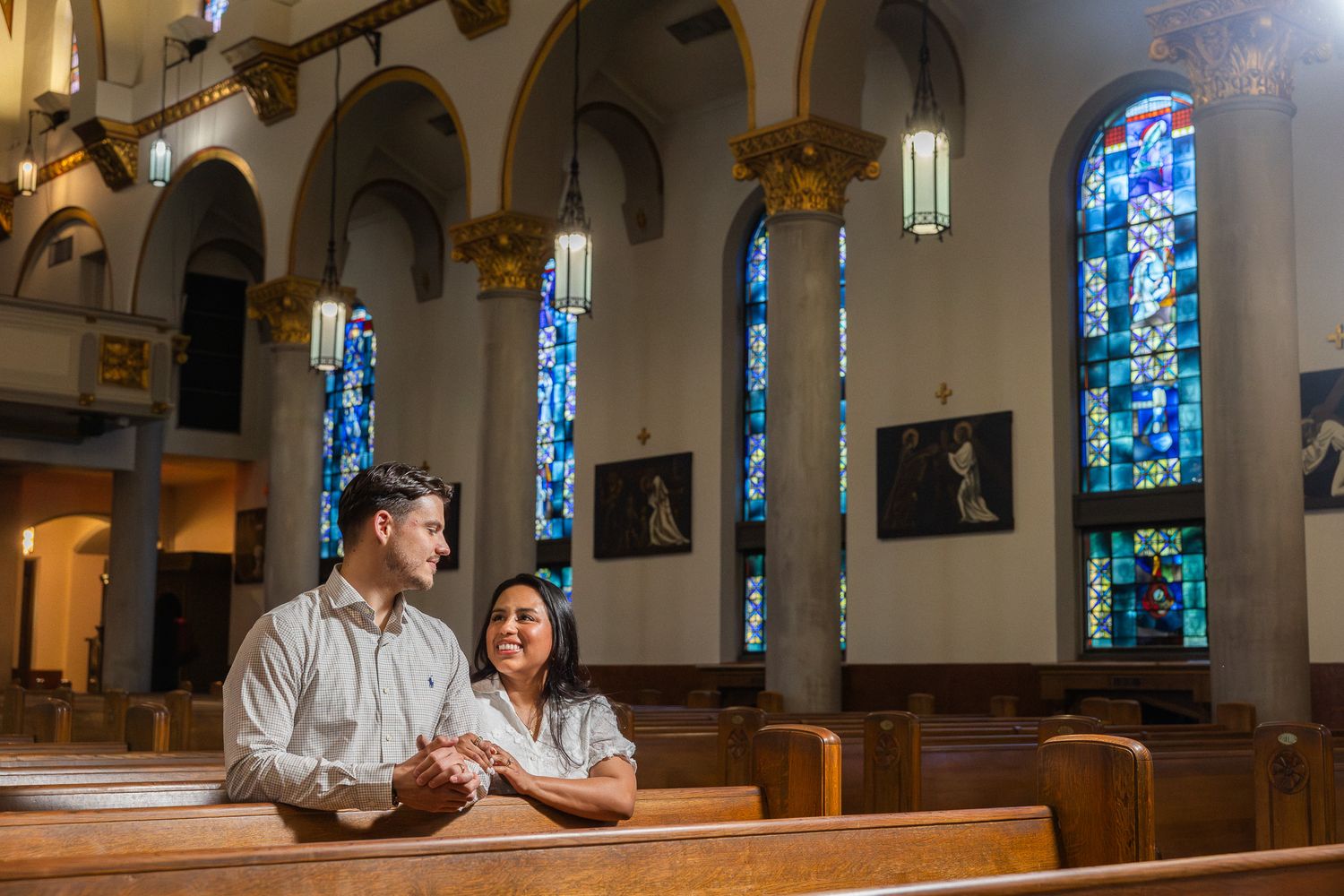 Couple sitting together in a church pew, smiling, with colorful stained glass windows in the background.