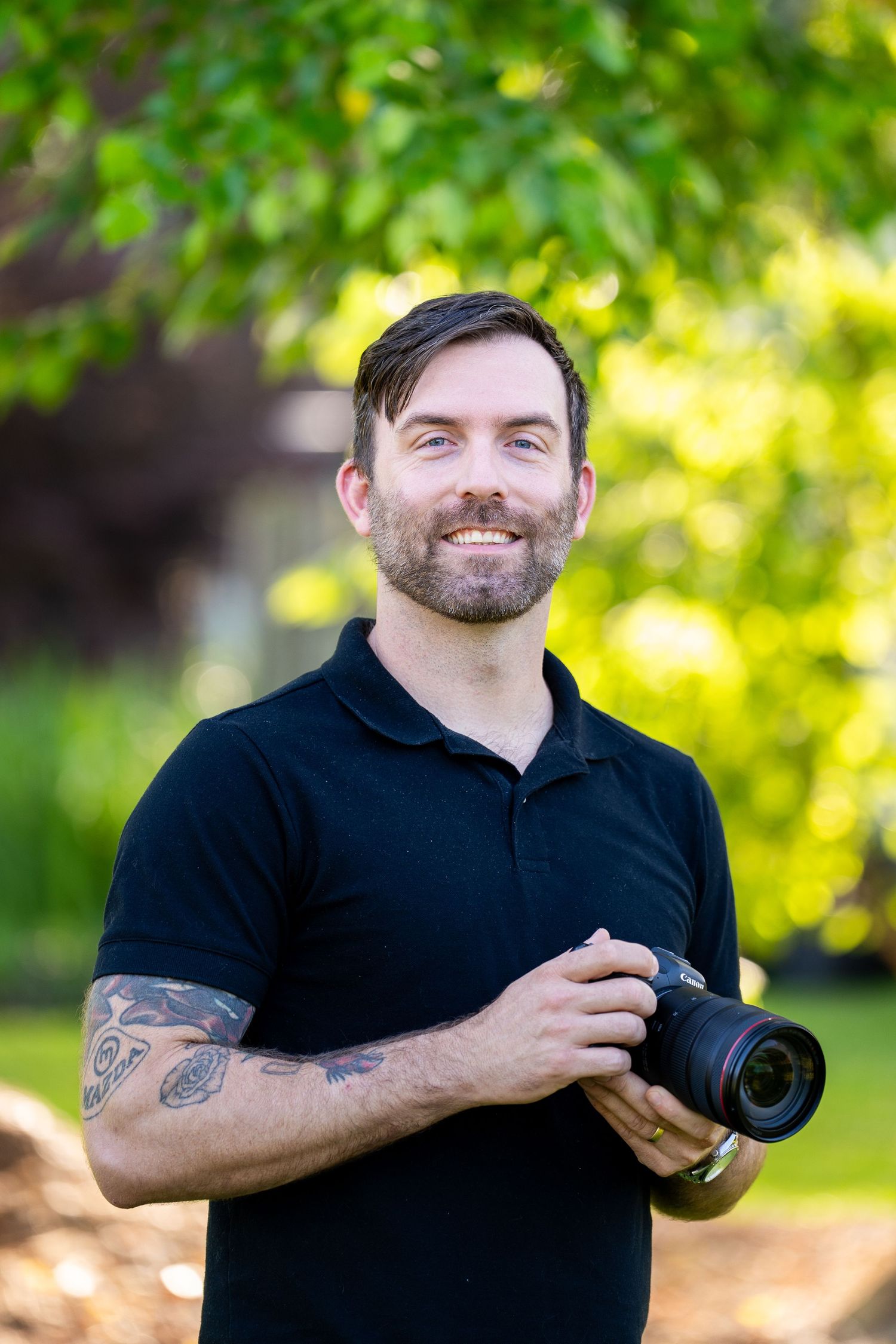 Smiling male photographer with tattoos holds DSLR camera outdoors among lush green trees.
