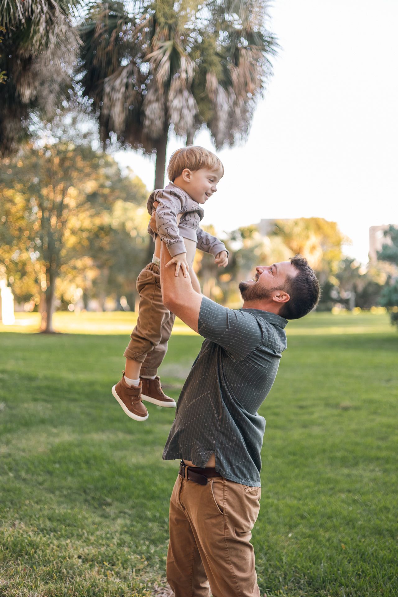 Father joyfully lifts toddler son in the air at a sunny green park with palm trees.