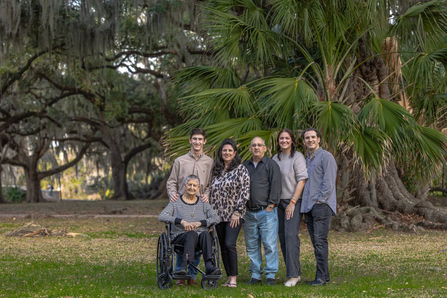 Family of six smiles together on green lawn with elderly woman in wheelchair beneath Spanish moss trees.
