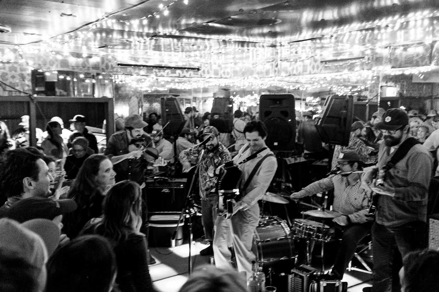 Live band performs energetically for a crowd at an indoor venue decorated with string lights.