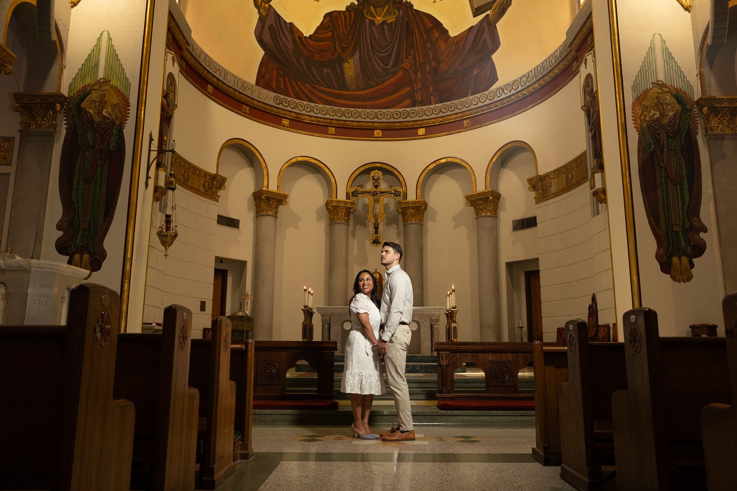 Couple poses together inside an ornate church with arched ceilings and a large religious mosaic mural above the altar.