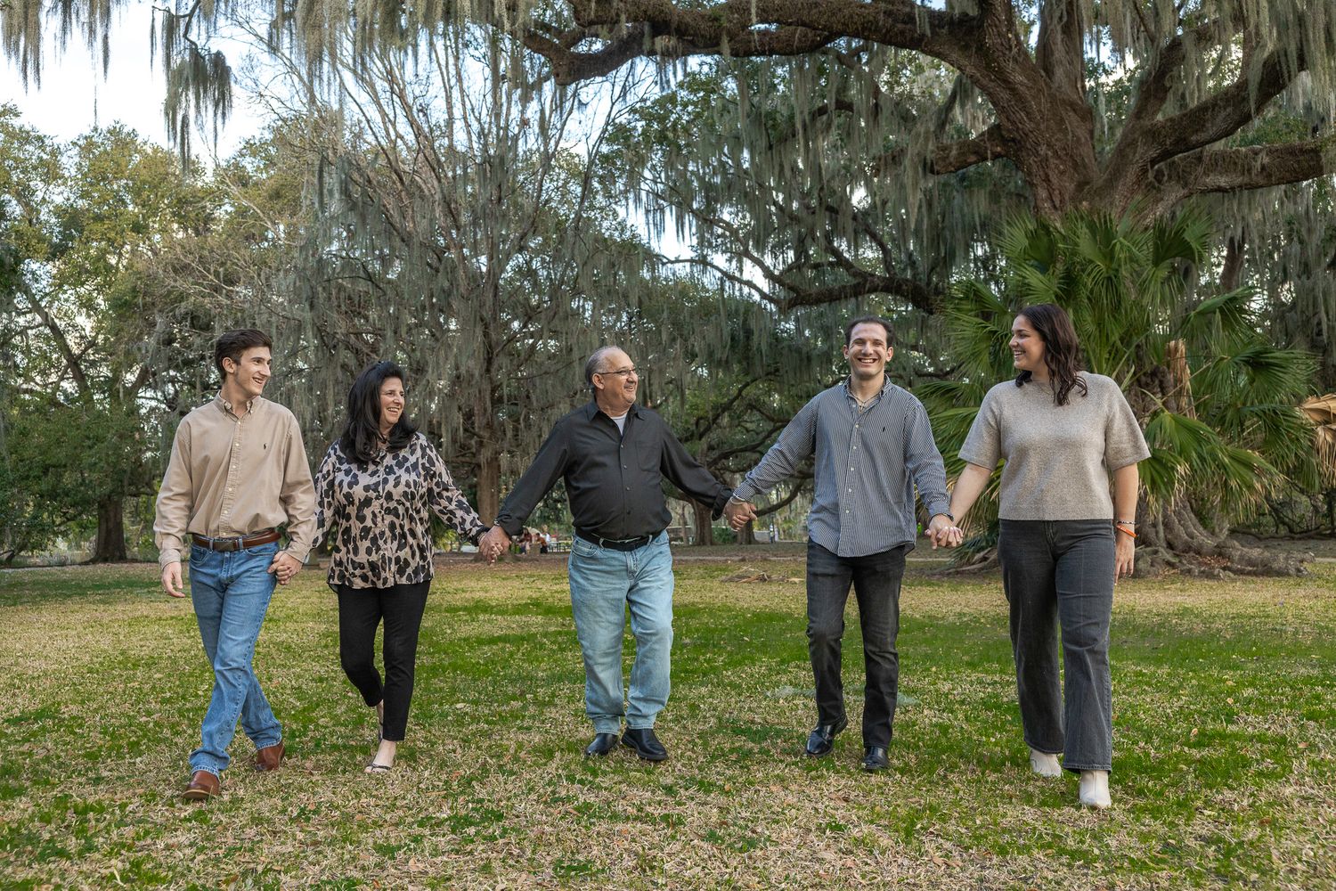 Five family members walk hand-in-hand across green lawn beneath large Spanish moss-covered oak trees.