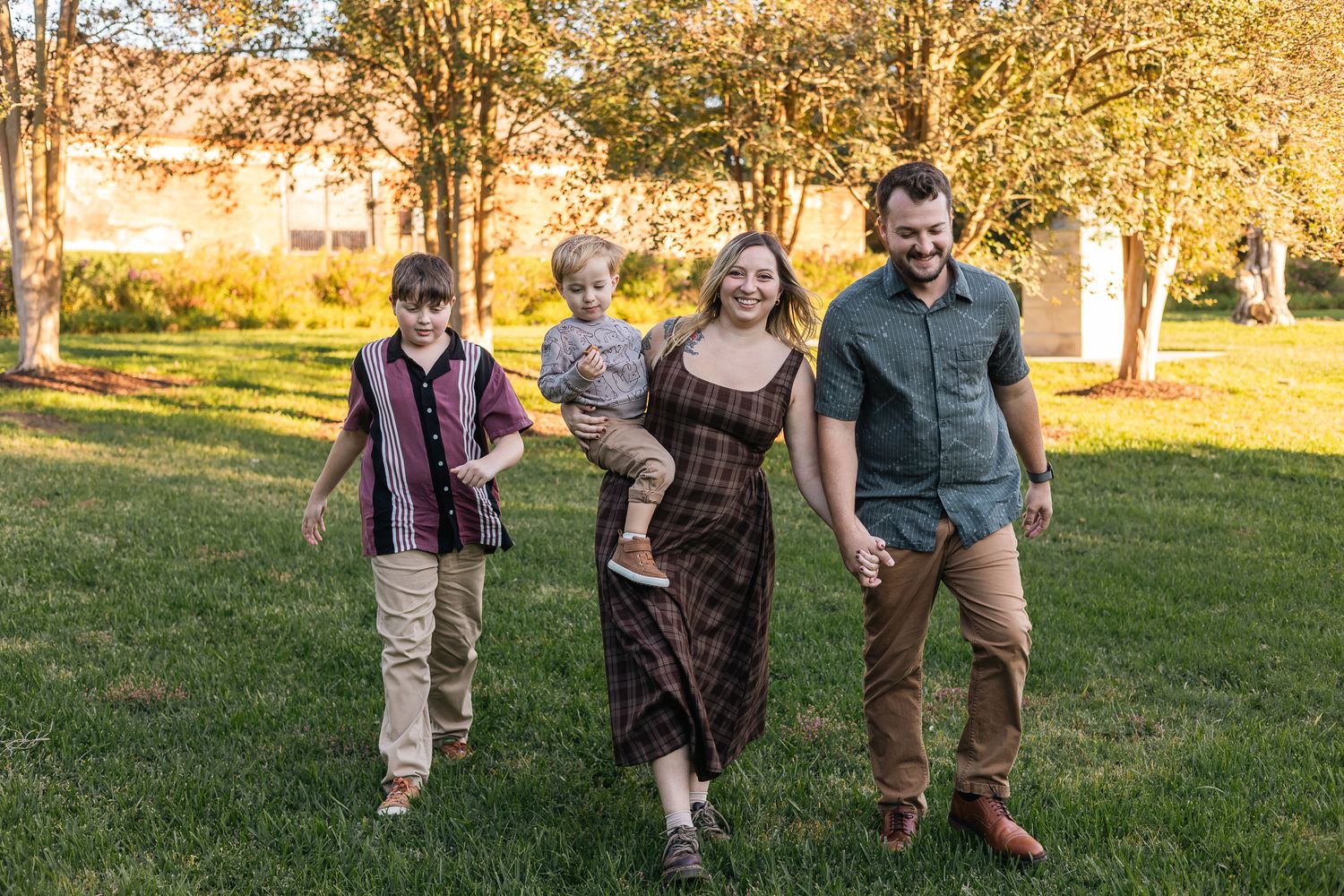 Happy family of four walks together on green grass during golden hour in autumn park setting.