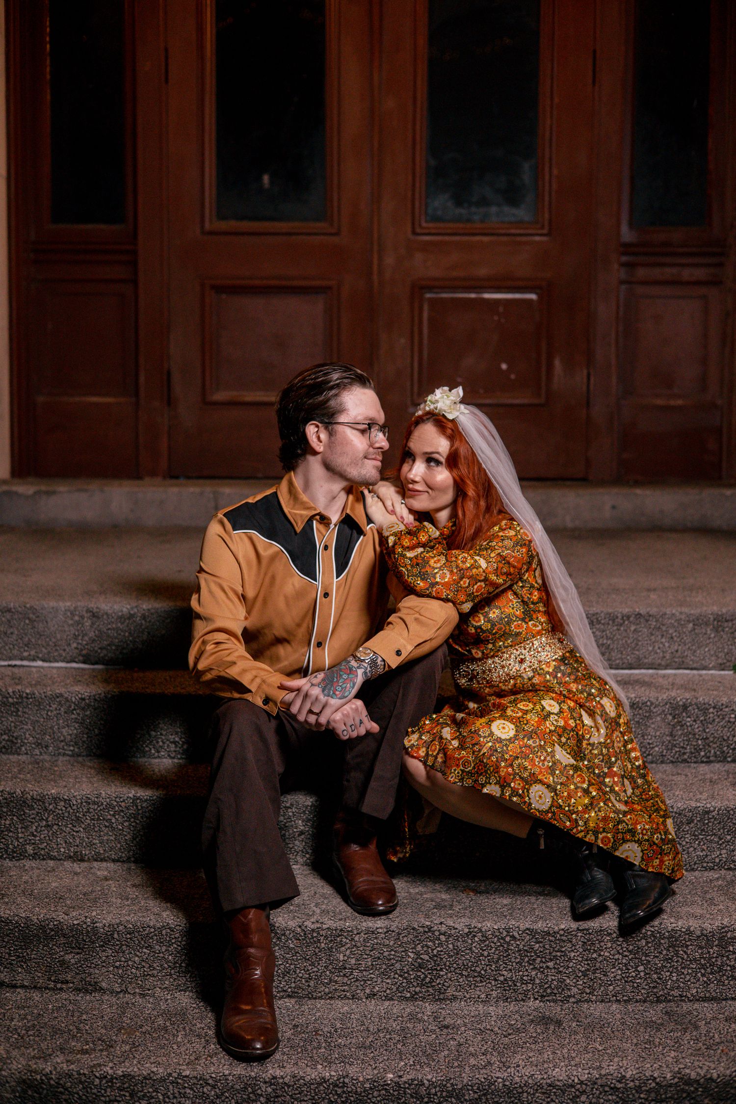 Couple in vintage 1970s attire sitting on steps, bride in floral dress with veil, groom in brown western shirt.