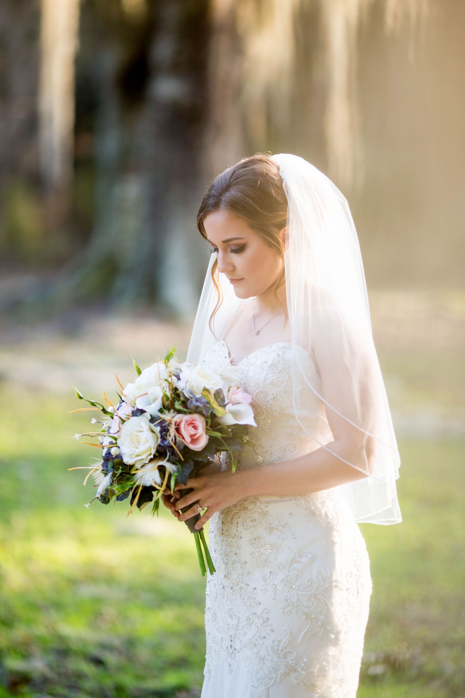 Bride in lace gown and veil gazes down at her pink and white floral bouquet.