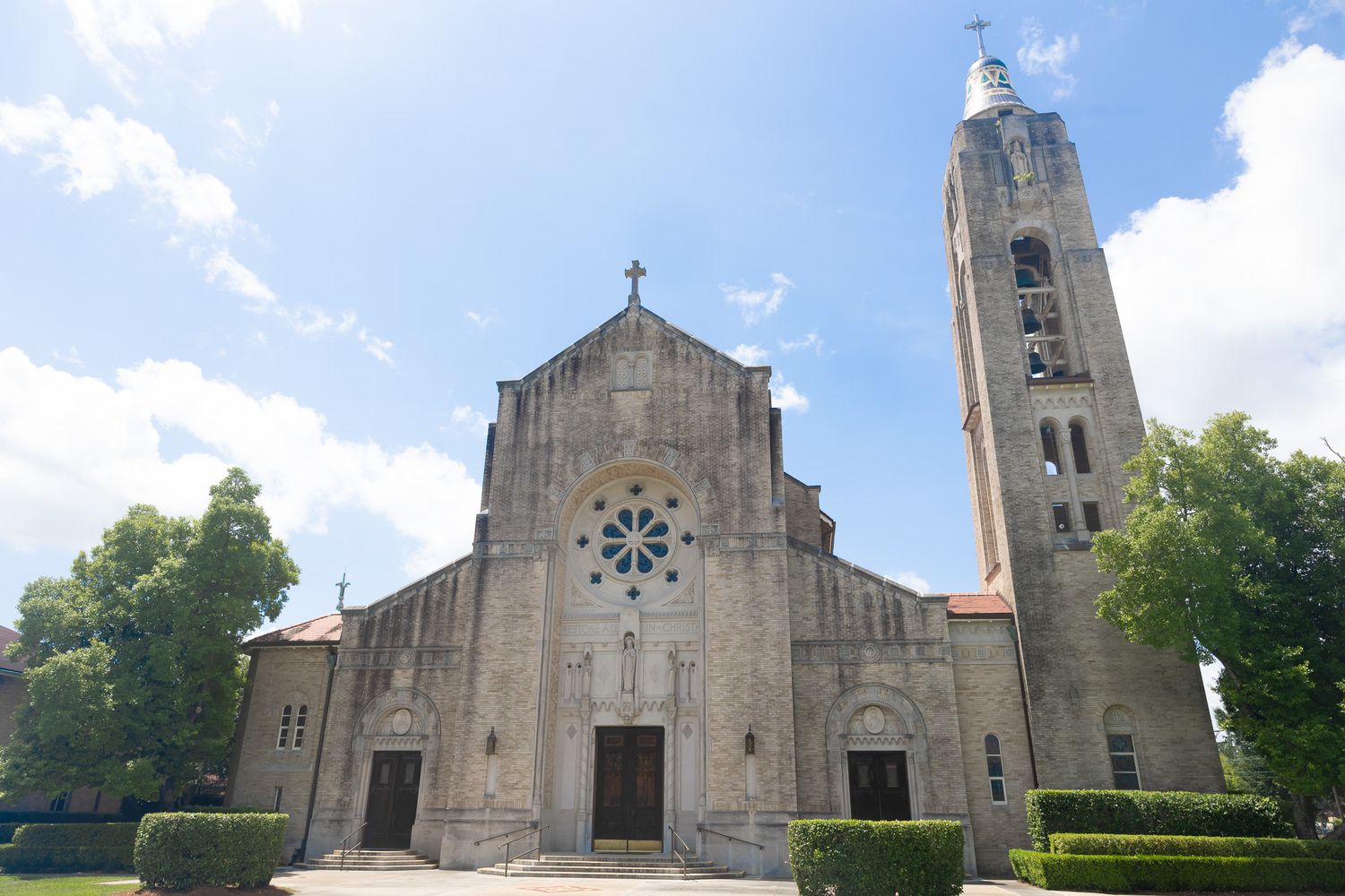 Historic stone Catholic church with rose window, tall bell tower, and manicured hedges under a blue sky.