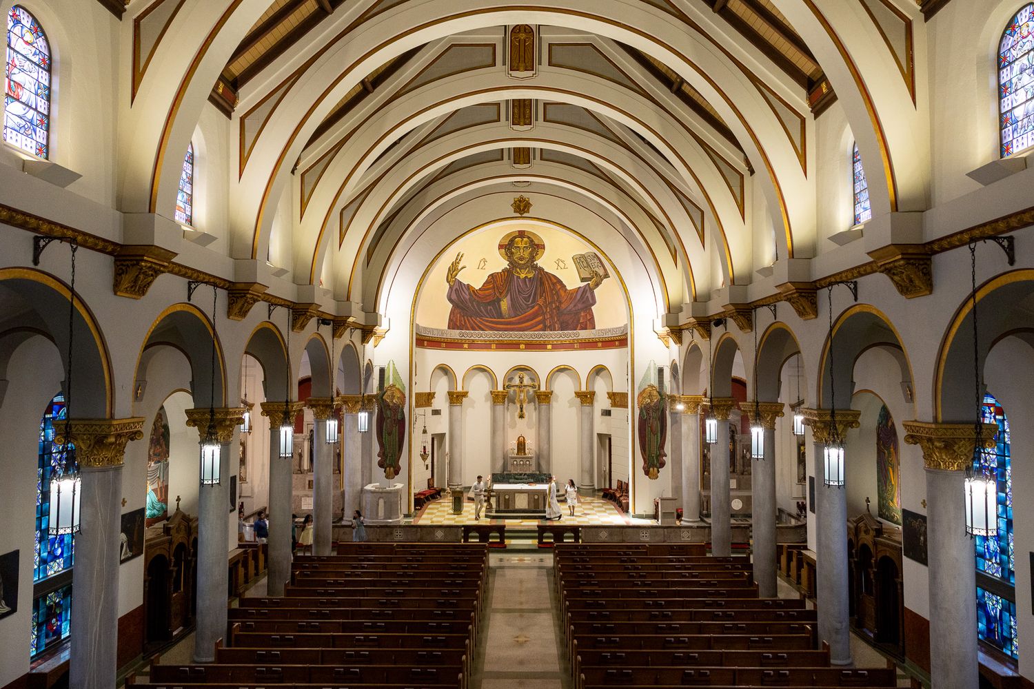 Interior of a grand Catholic church featuring wooden pews, arched ceilings, stained glass windows, and a mosaic of Christ above the altar.