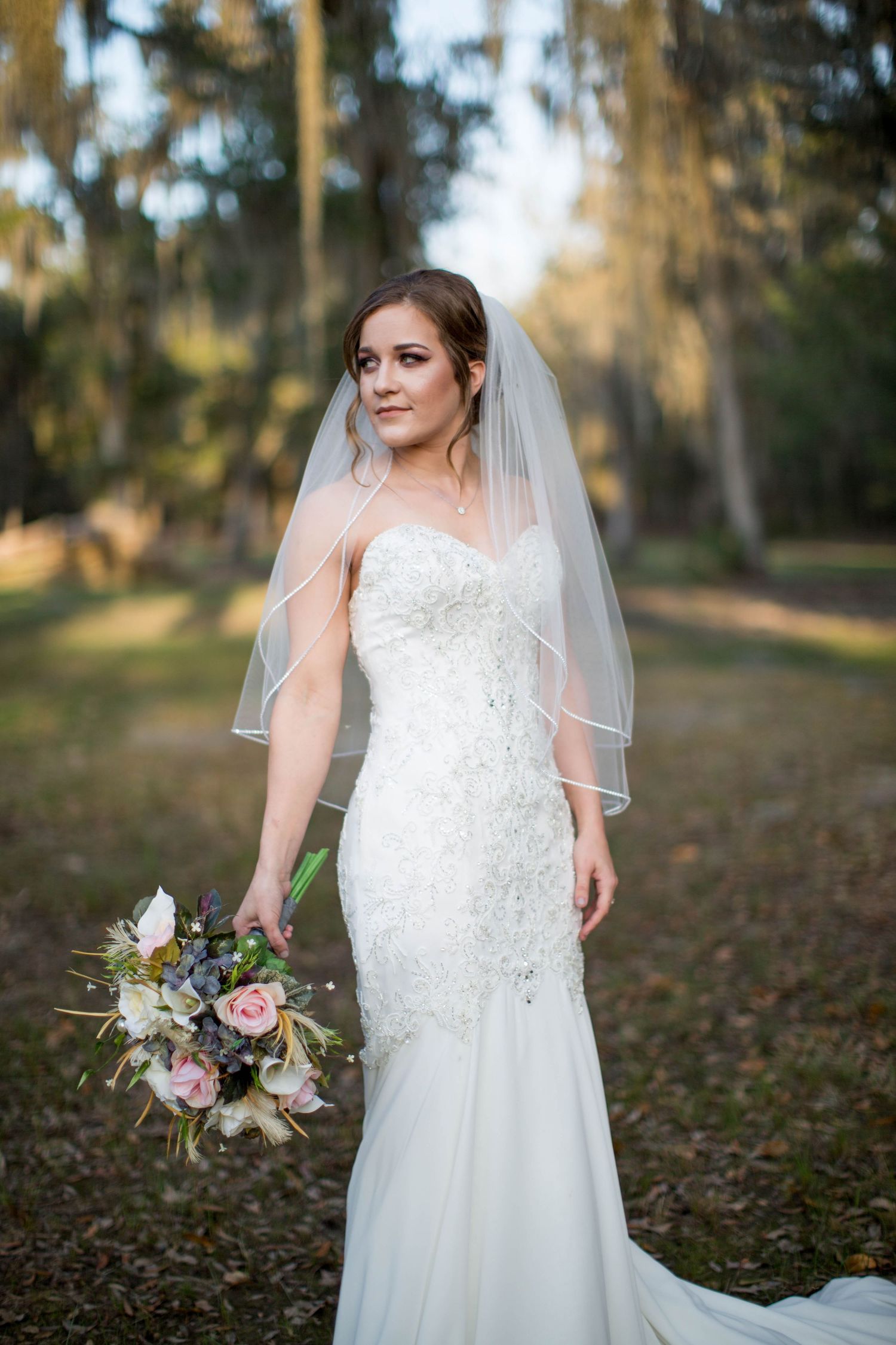 Bride in white lace mermaid gown with veil holds floral bouquet outdoors among trees.