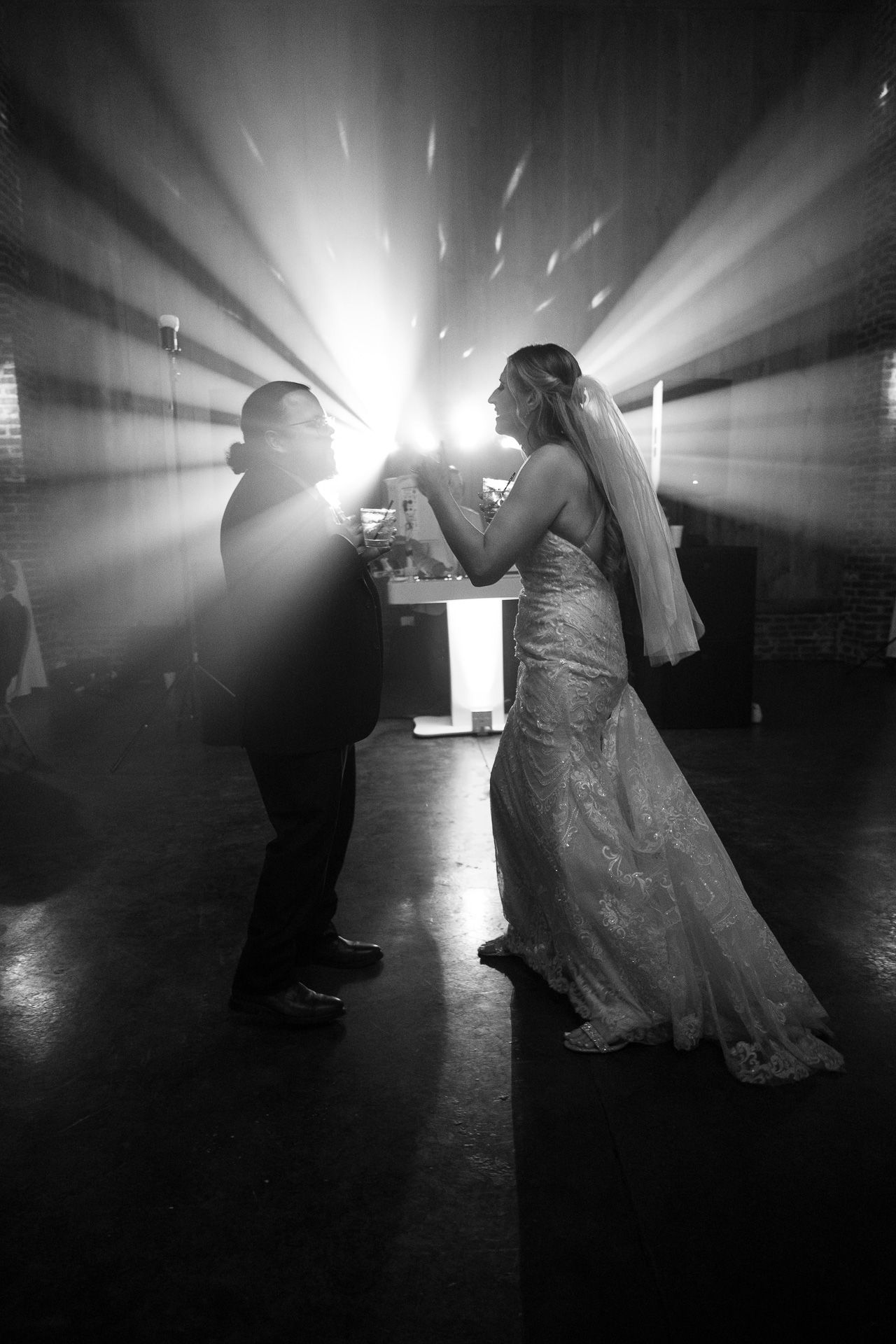 Black and white photo of bride and groom sharing first dance with dramatic light rays behind them.