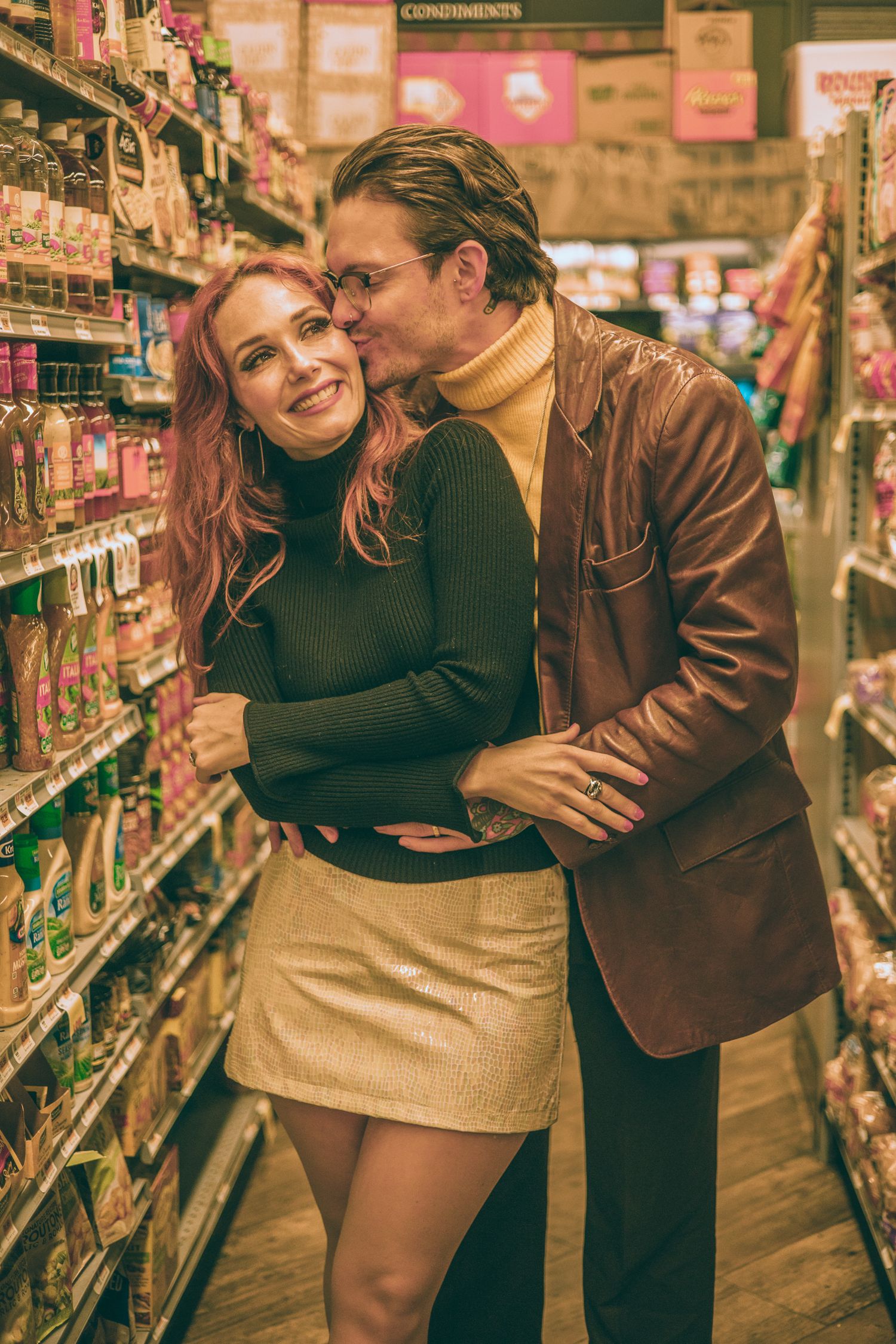 A couple shares a romantic embrace in a grocery store aisle, woman in mini skirt smiling as man kisses her cheek.