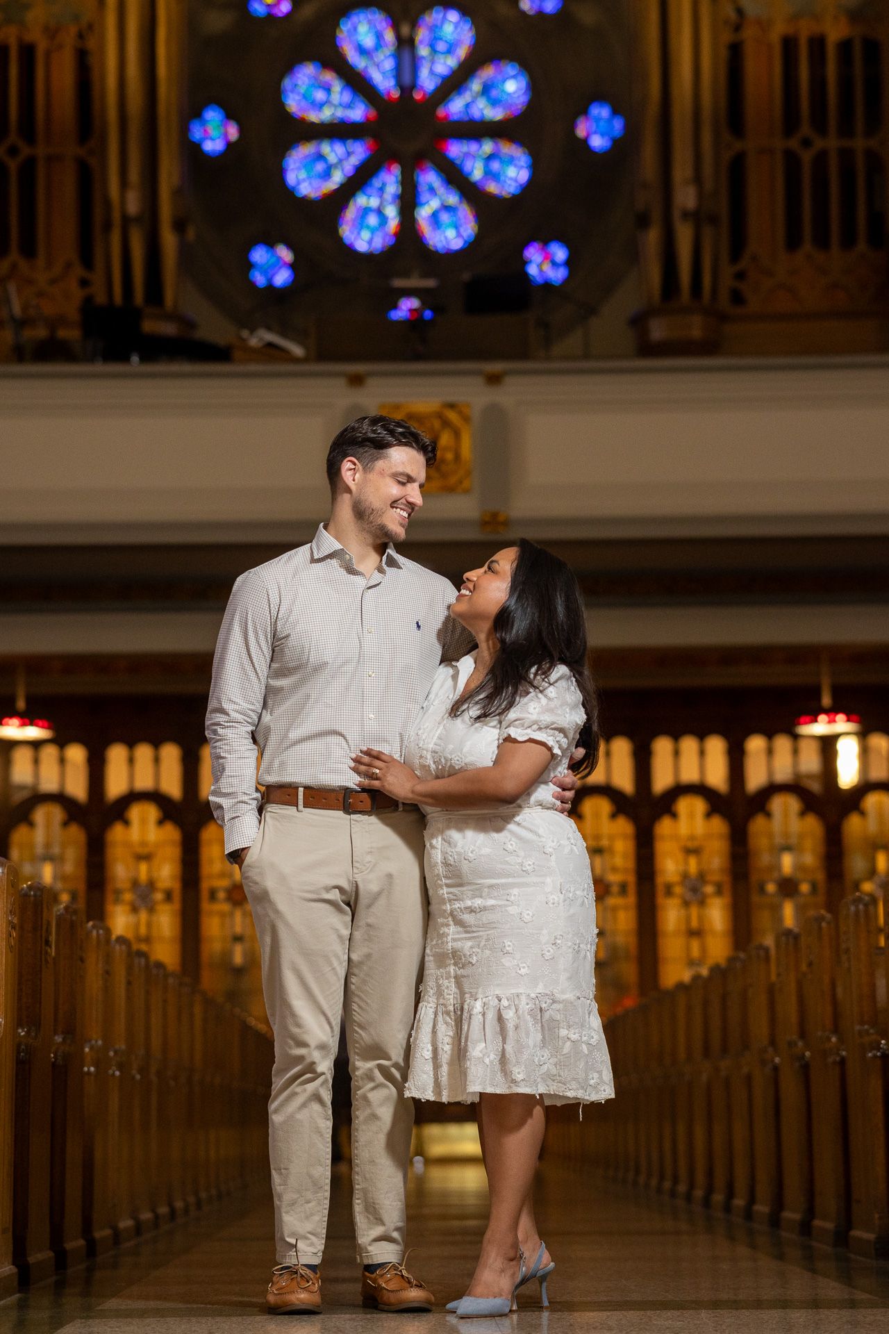 Couple embracing in church aisle with stunning stained glass rose window backdrop, wearing white outfits.