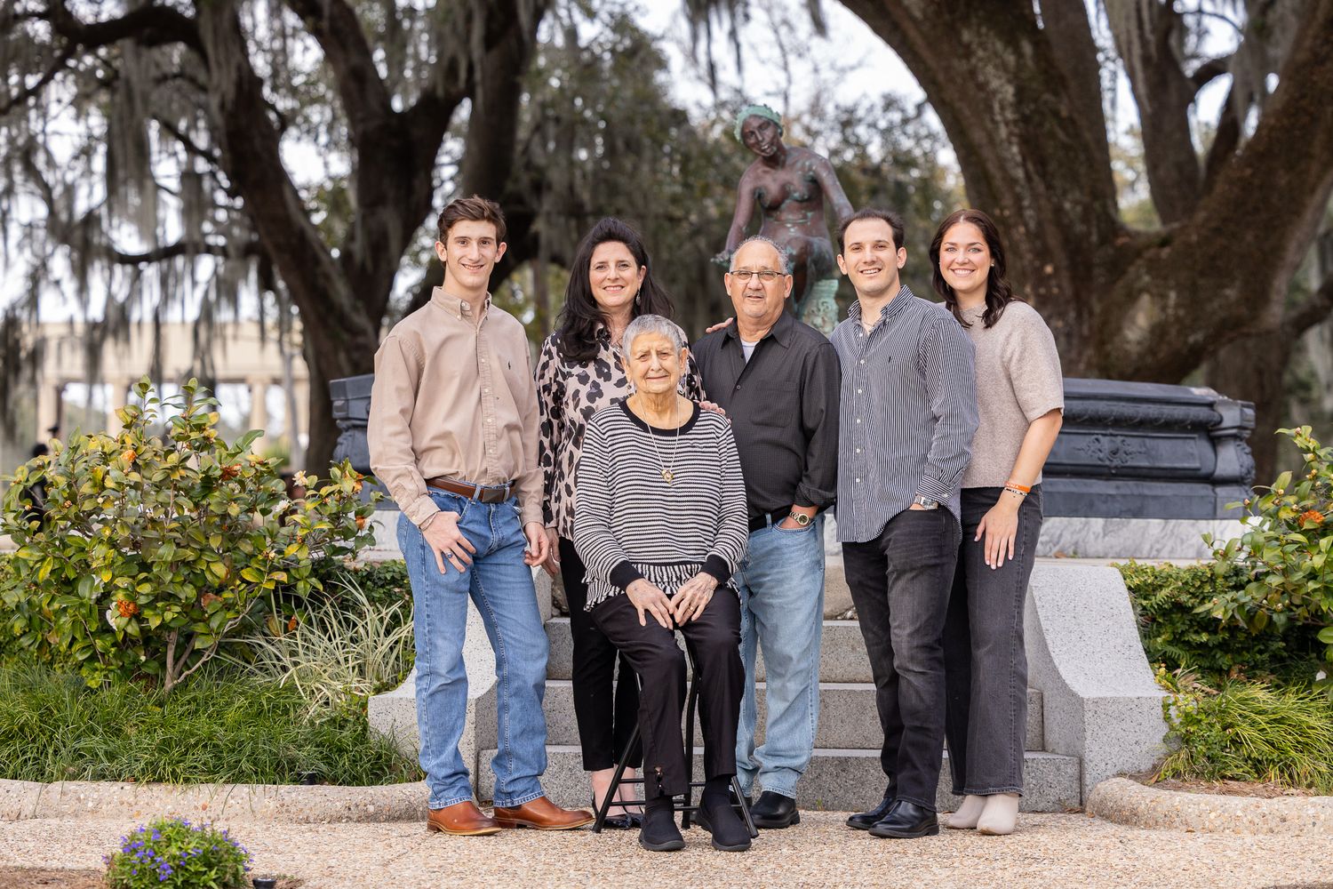 Six family members pose together near bronze statue under Spanish moss-draped oak trees in historic park.