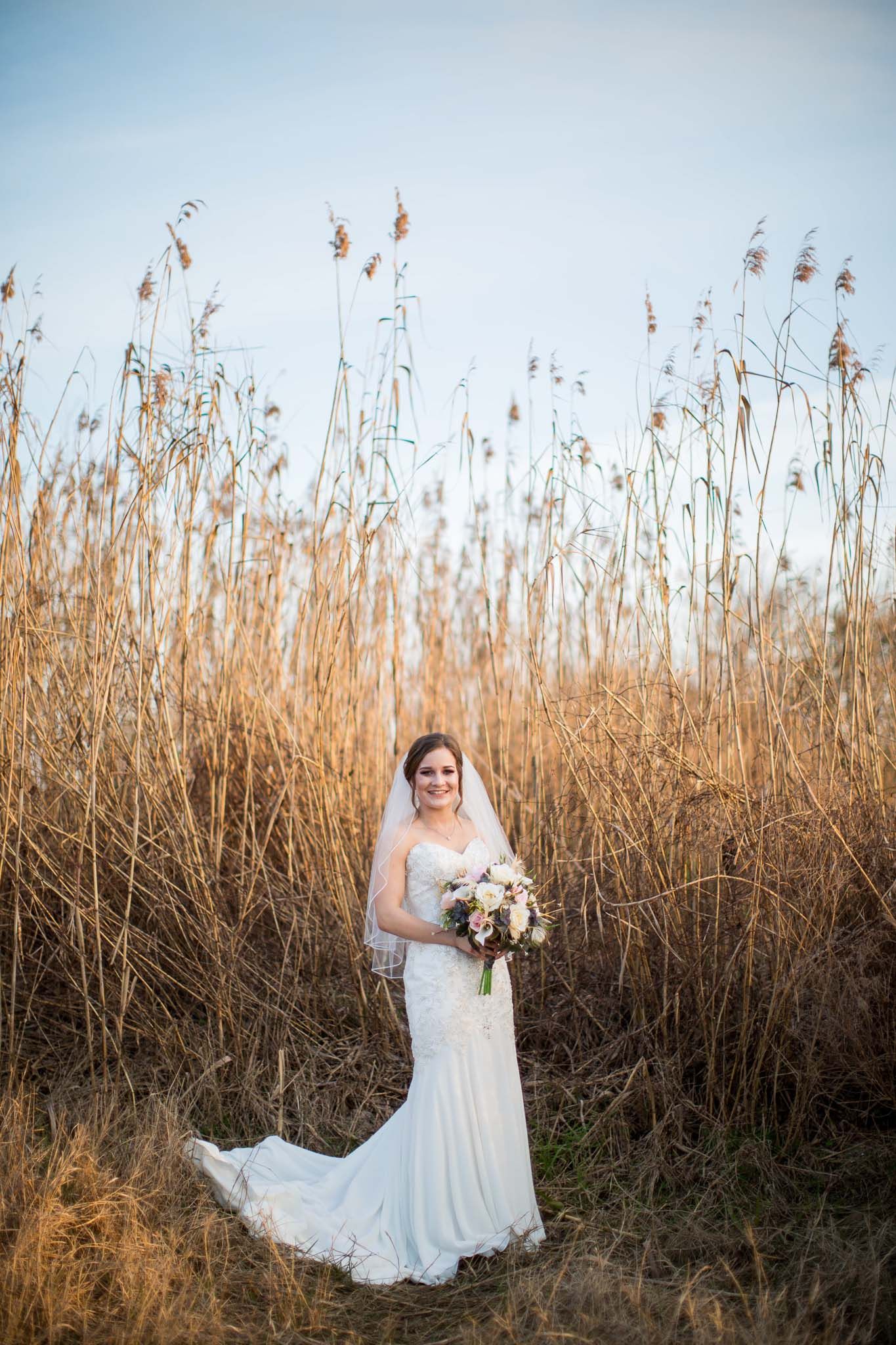 Bride in white lace gown holds bouquet while standing in golden tall grass field.