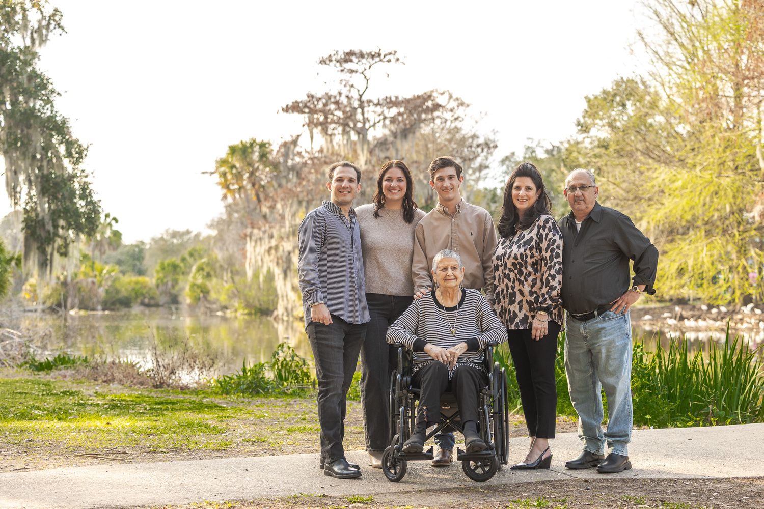 Multi-generational family portrait with grandmother in wheelchair along scenic waterway path in sunny park.