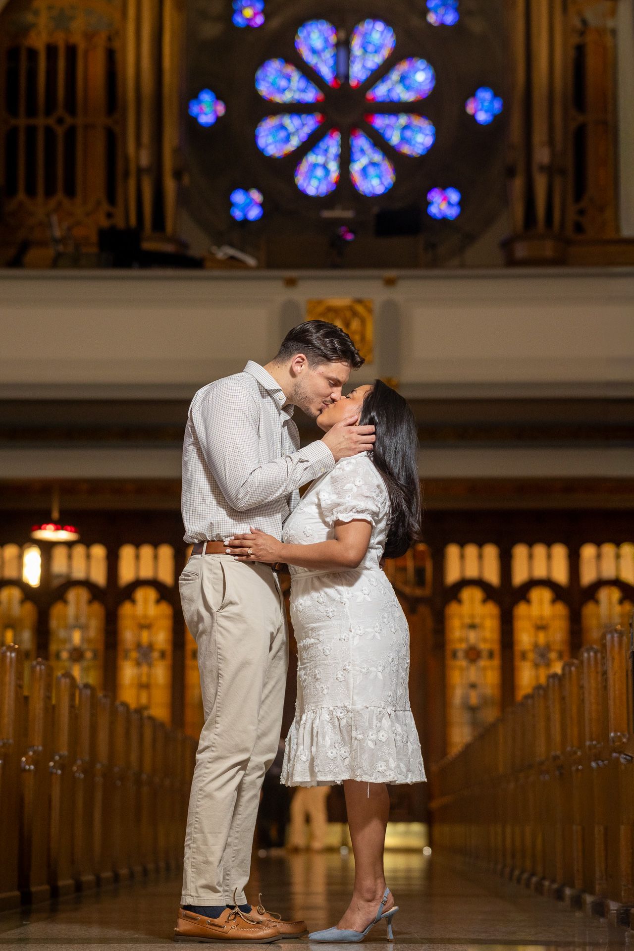 Couple sharing a romantic kiss inside a church with wooden pews and a stunning stained glass rose window backdrop.