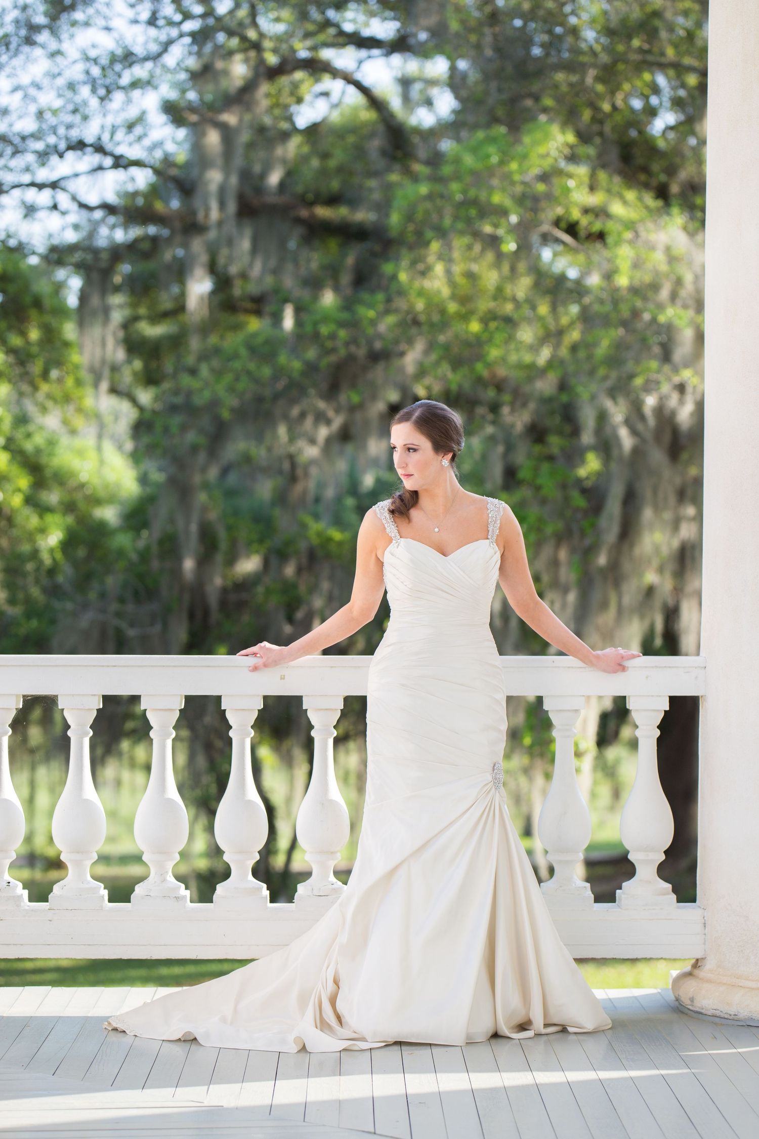Elegant bride in white mermaid gown poses on a white balcony surrounded by lush trees.