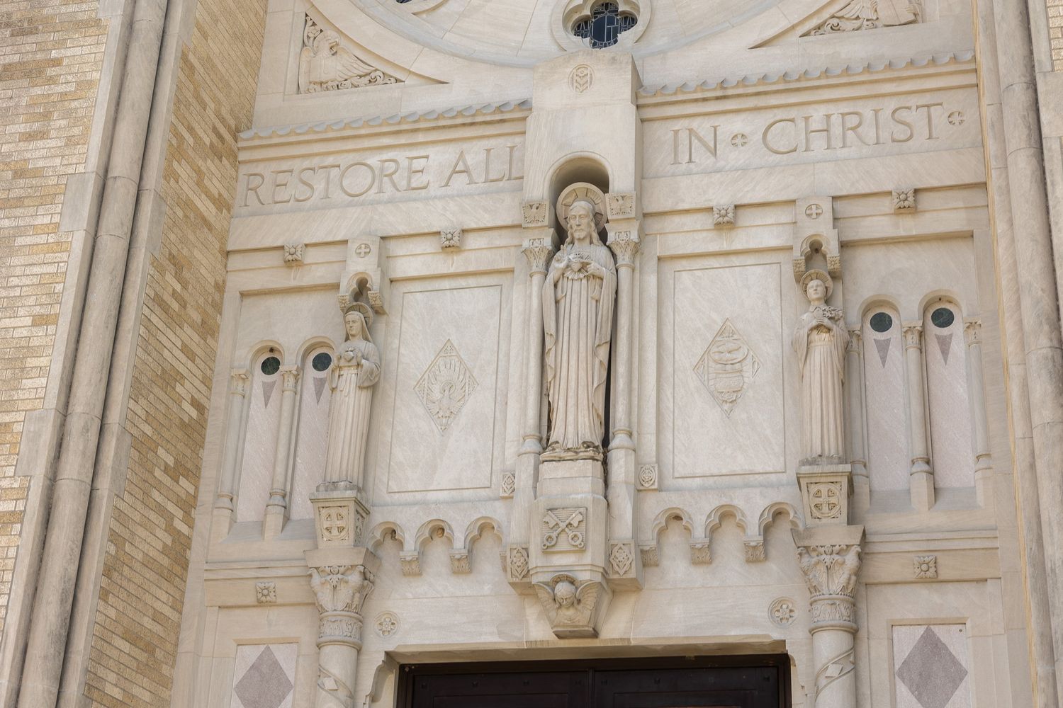 Stone church facade with statue of Virgin Mary and inscription 'Restore All In Christ' above ornate entrance doors.