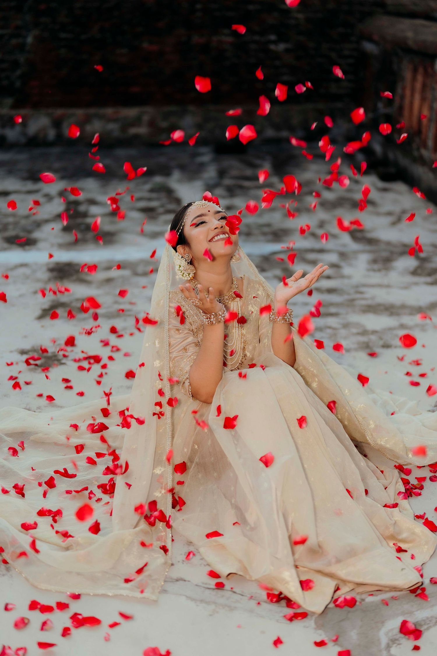 Indian bride in cream lehenga joyfully catching falling red rose petals at wedding celebration.