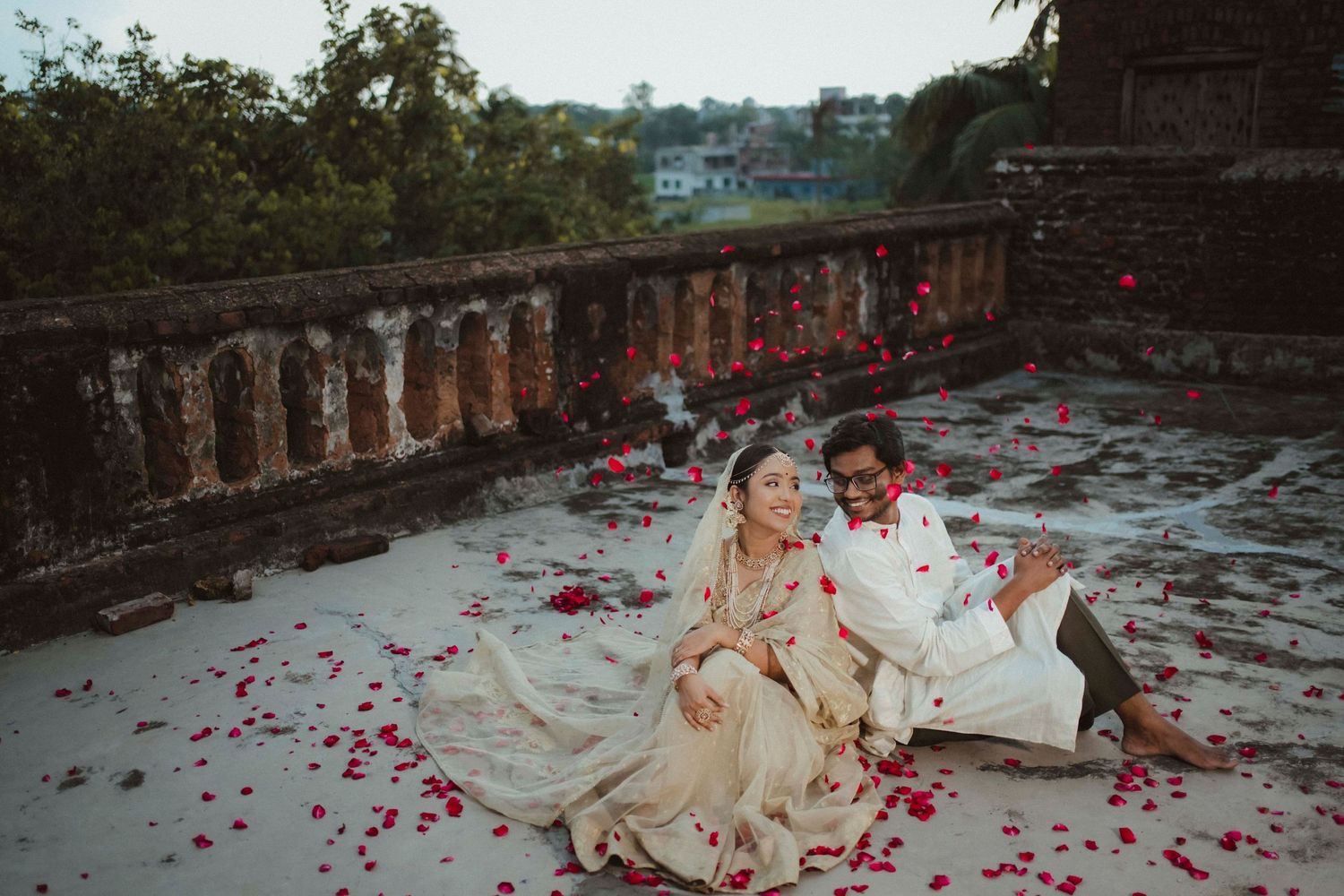 Indian wedding couple sitting together on rooftop surrounded by scattered red rose petals at dusk.