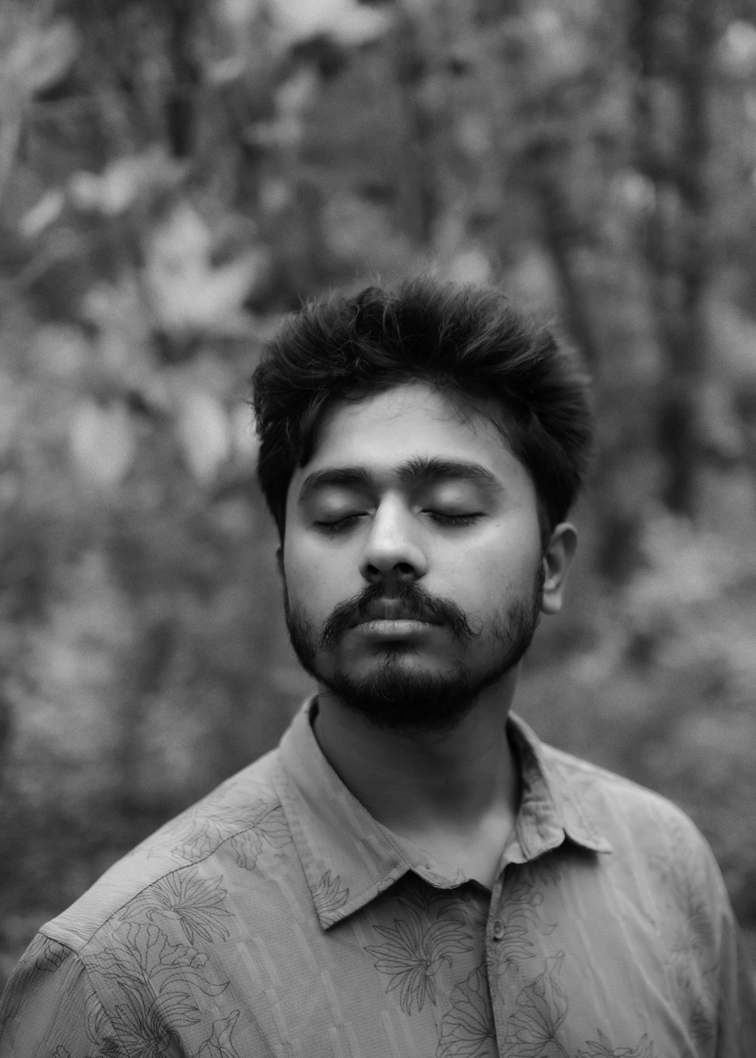 Black and white close-up portrait of a young bearded man with closed eyes in a floral shirt outdoors.