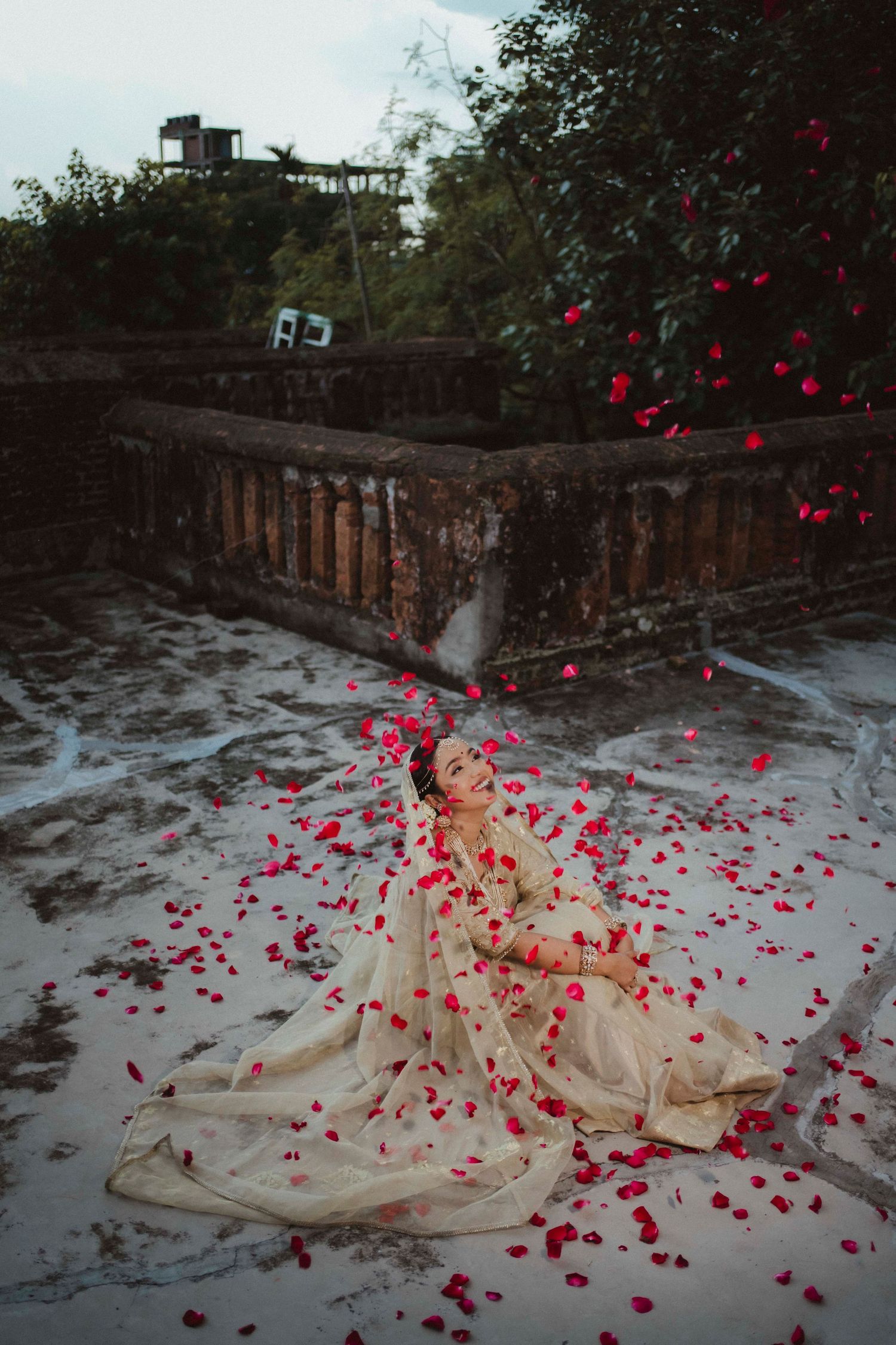 Indian bride in ivory bridal lehenga sitting on rooftop as red rose petals rain down around her.