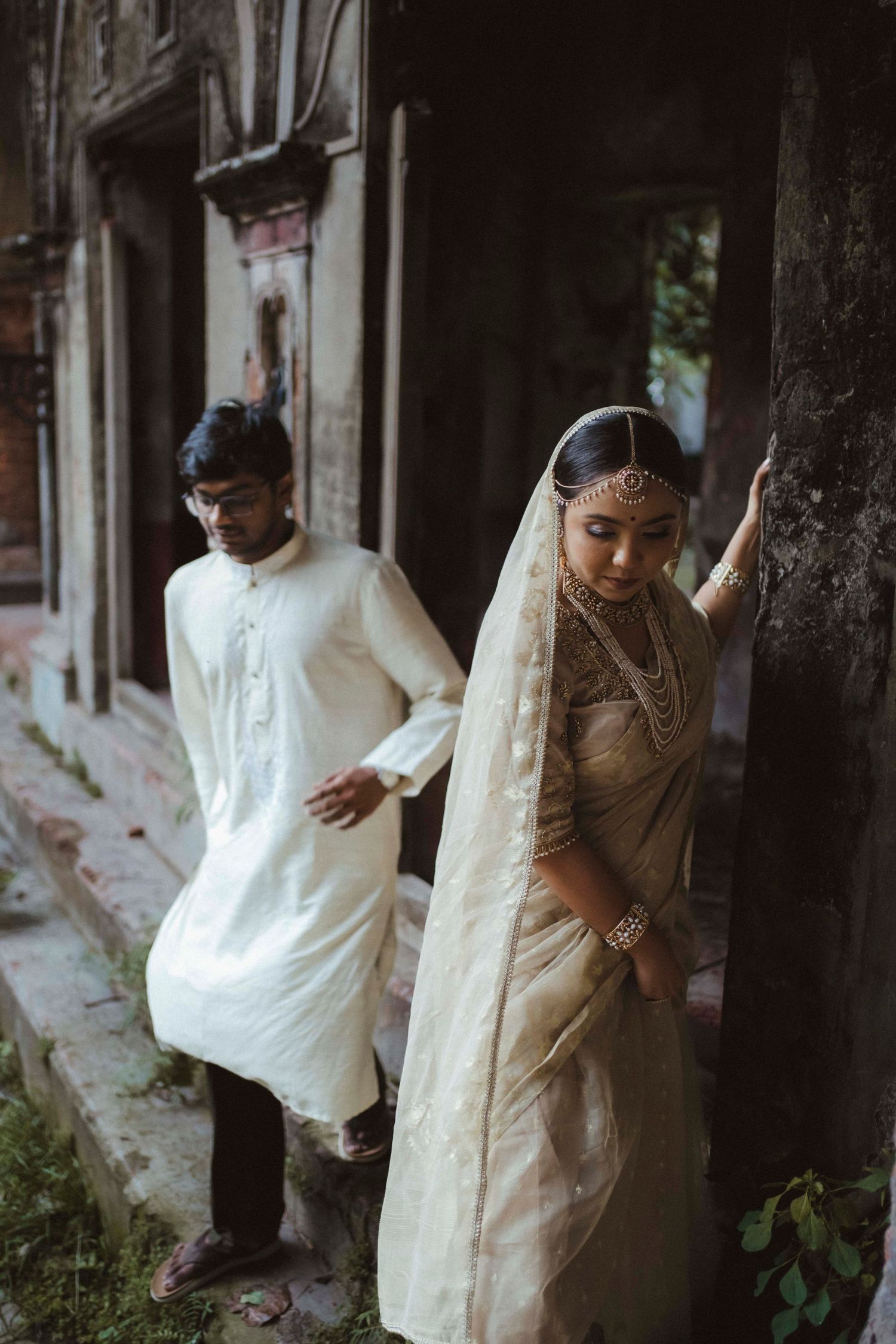Indian wedding couple walking through rustic heritage building corridor in traditional cream bridal attire.