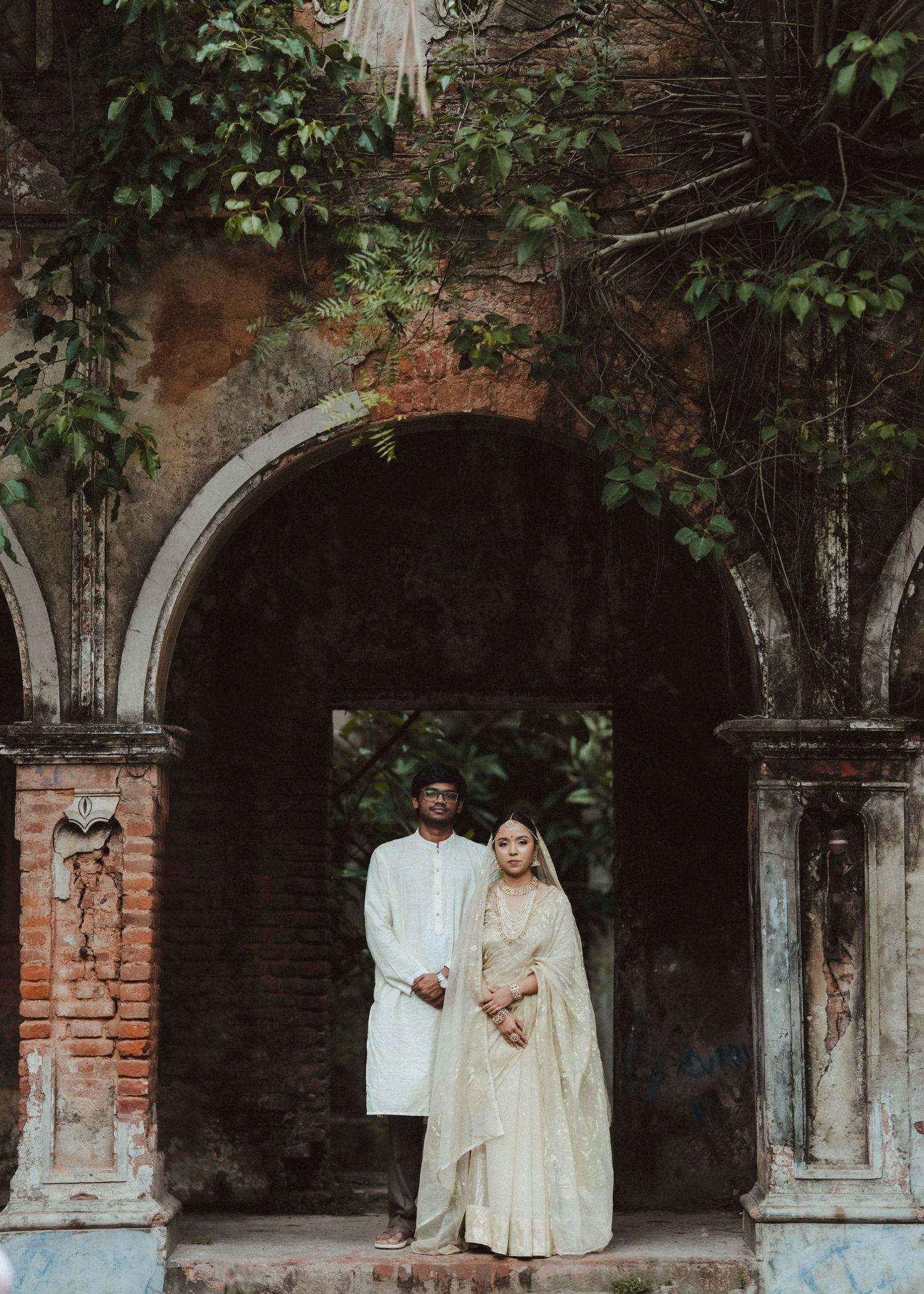Indian wedding couple standing under ornate brick archway surrounded by lush green foliage.