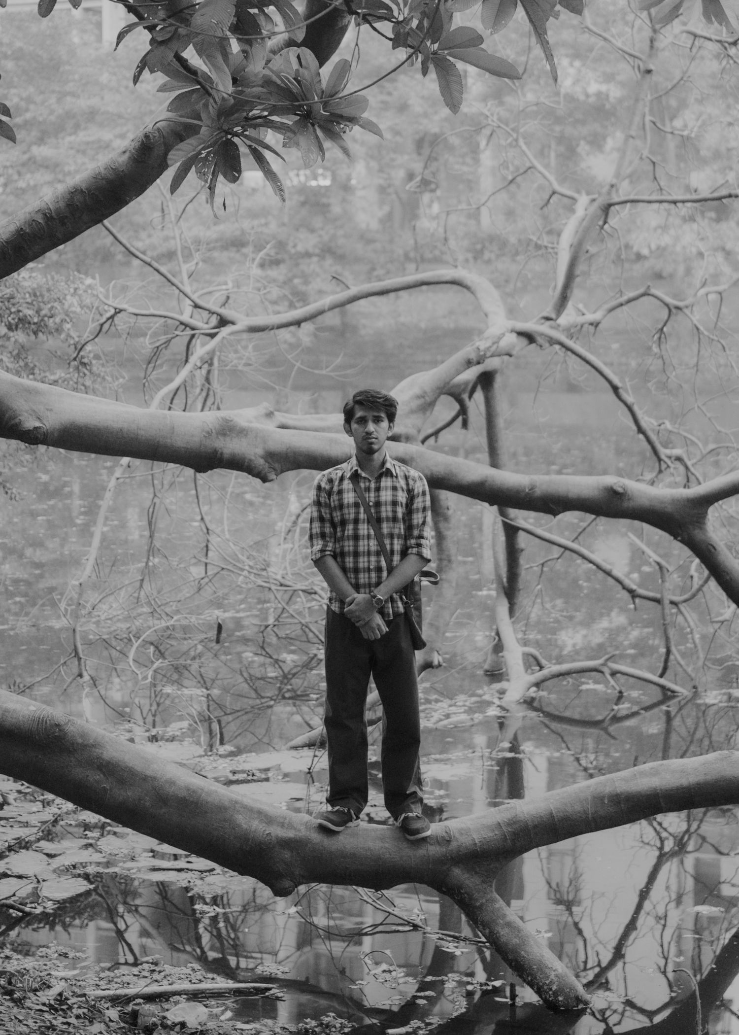 Black and white photo of a young man standing on large tree branches near a reflective water body.