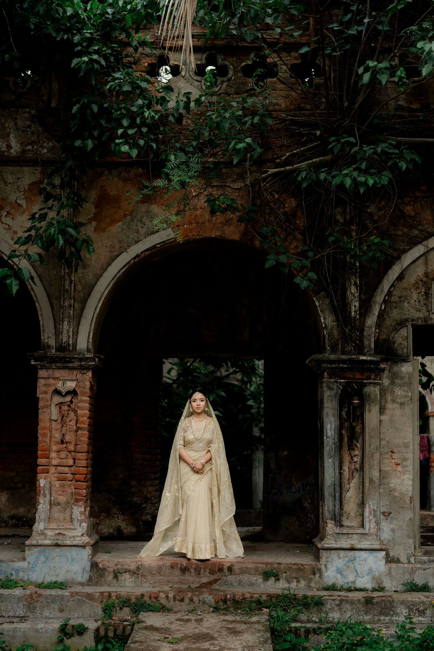 A bride in cream lehenga and dupatta stands in an ancient arched ruin surrounded by lush greenery.