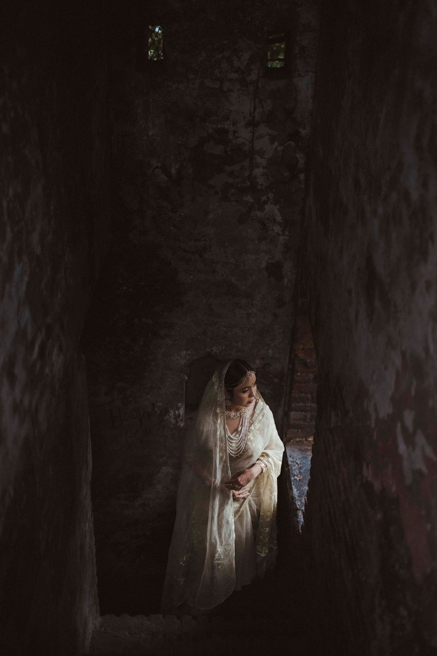 Indian bride in cream bridal saree standing in dramatic shadowy stone corridor of heritage building.