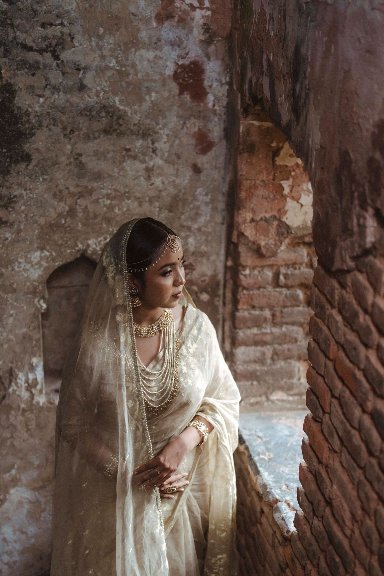 Indian bride in ivory saree adorned with gold jewelry posing in ancient brick archway.