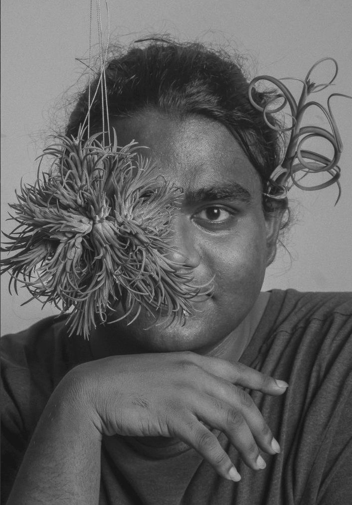 Black and white portrait of a woman holding a dried flower over half her face with curly plant stems in her hair.