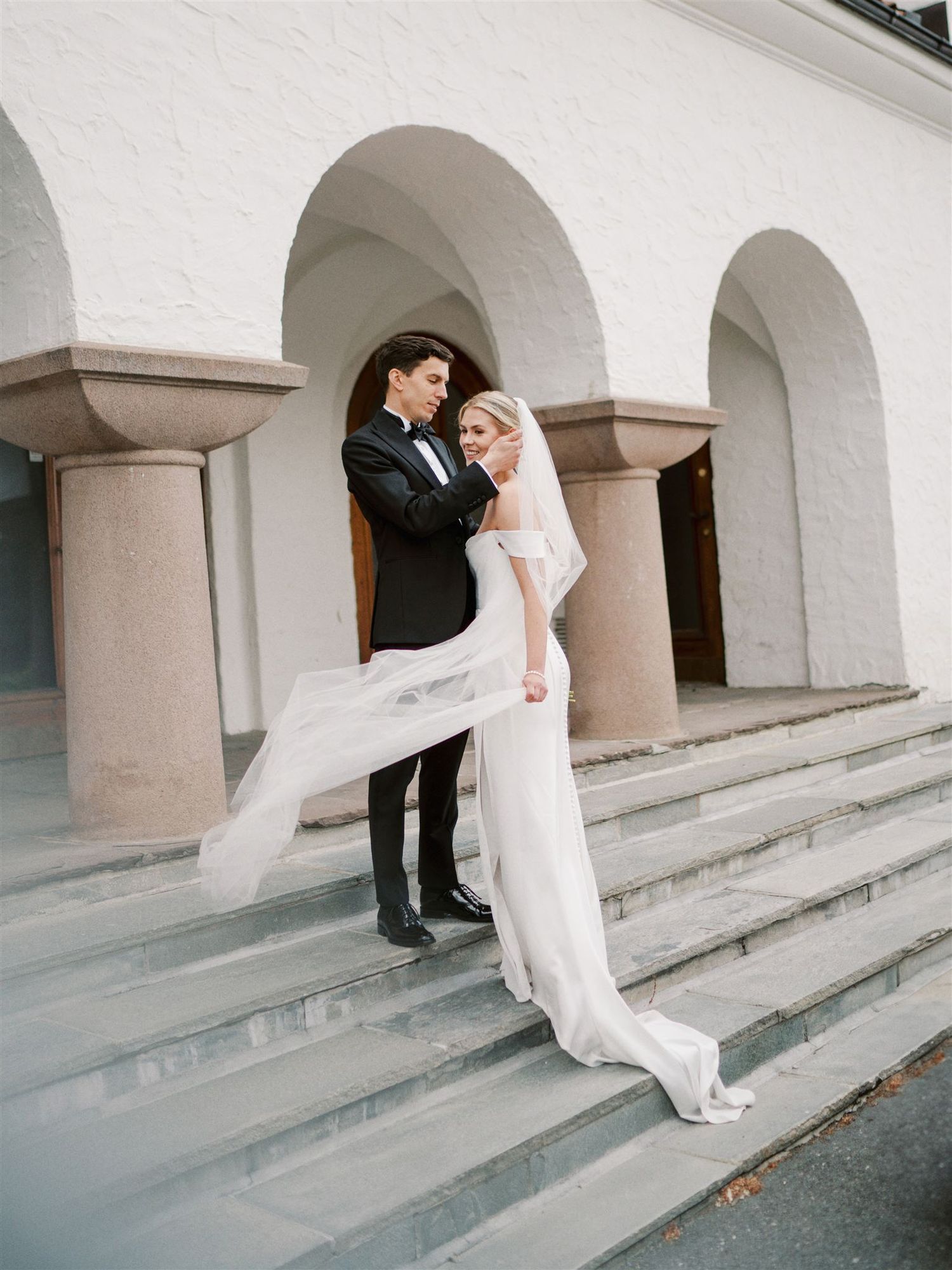 Bride and groom stand on stone steps of white arched building, veil flowing in the breeze.