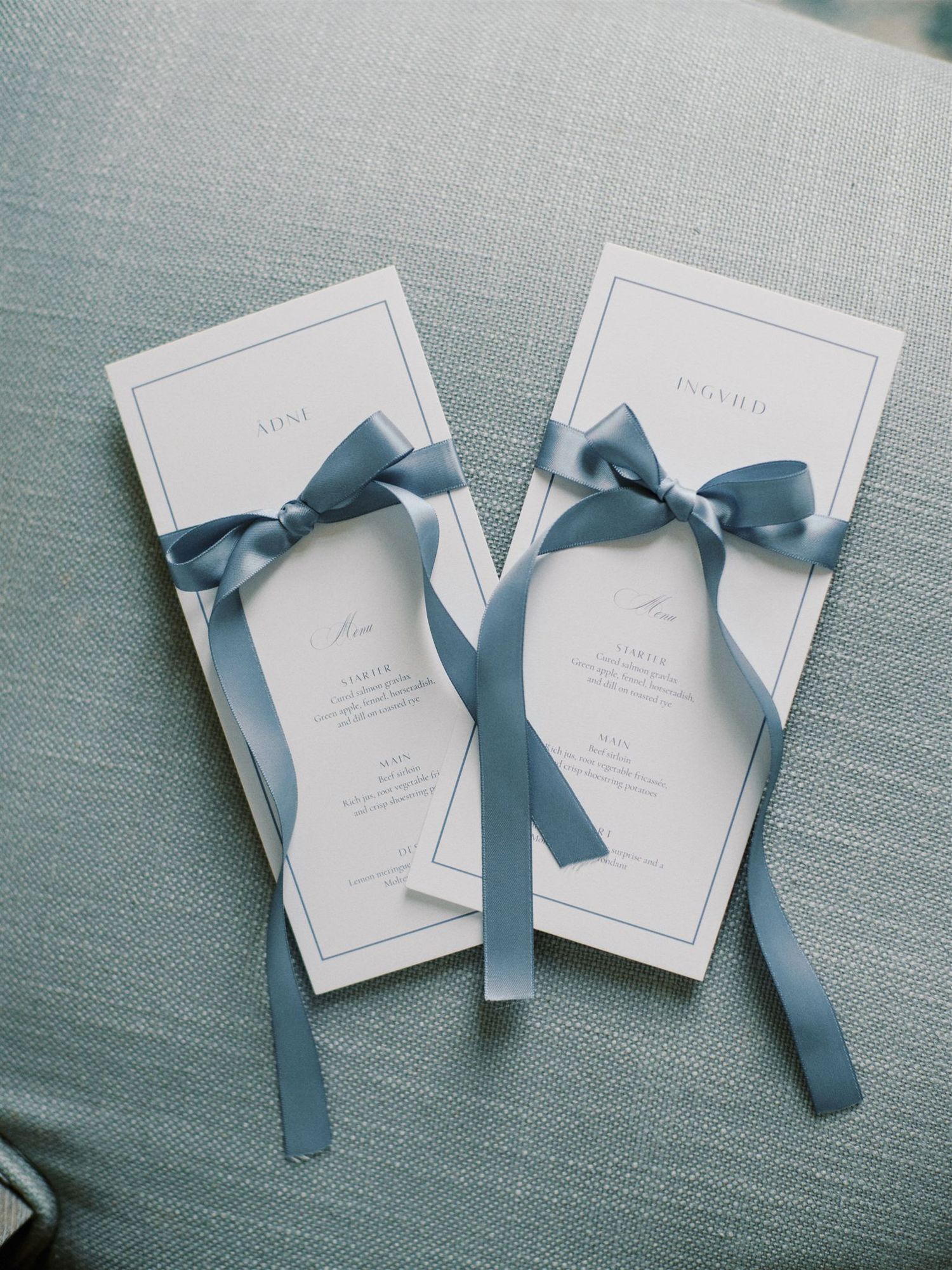 Elegant wedding programs with dusty blue satin ribbon bows resting on light blue fabric surface.