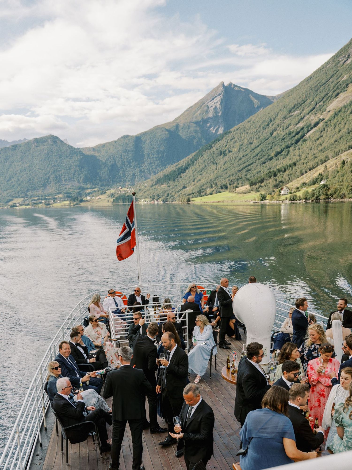 Elegant wedding guests gather on a boat deck with Norwegian flag, surrounded by stunning fjord and mountain scenery.