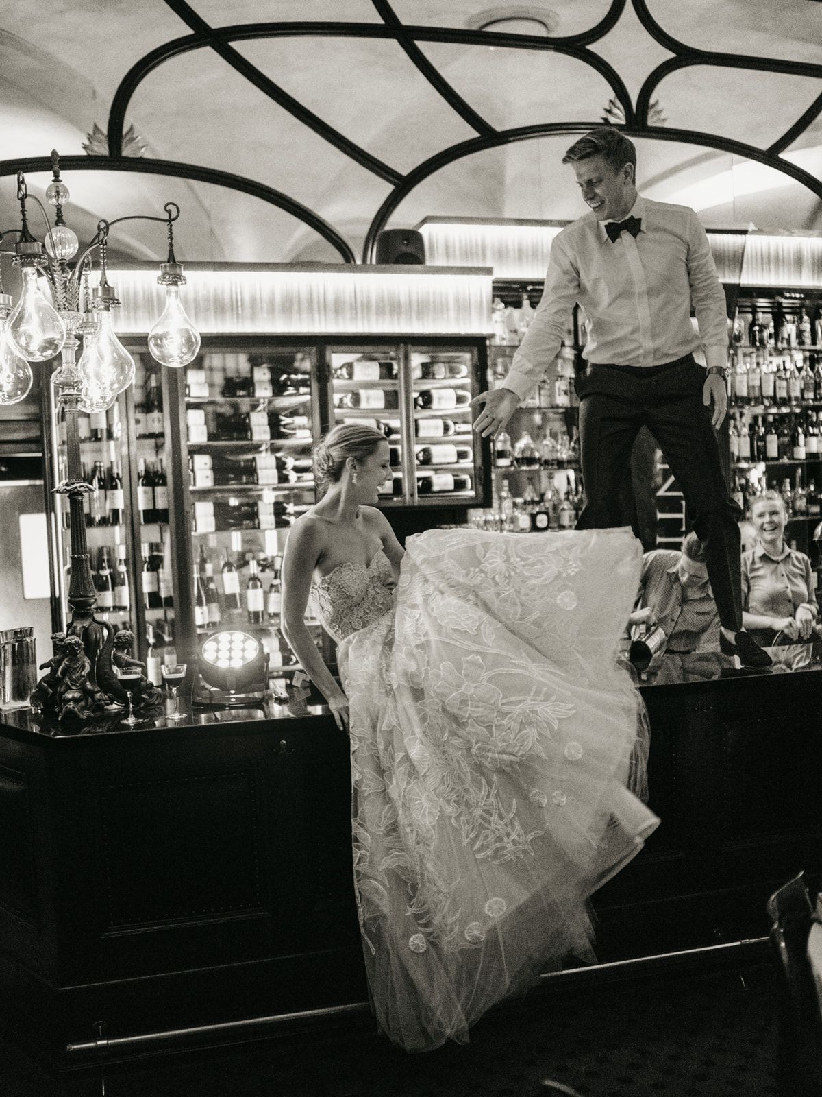 Bride in lace gown and groom standing on a bar counter in a classic black and white photo.