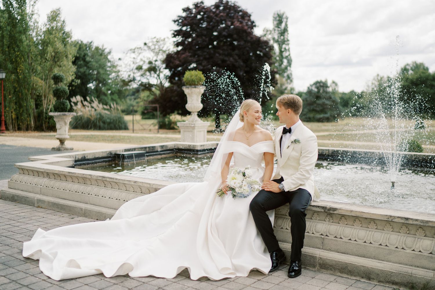 Bride and groom sitting by a stone fountain in lush garden, bride in flowing cathedral gown.