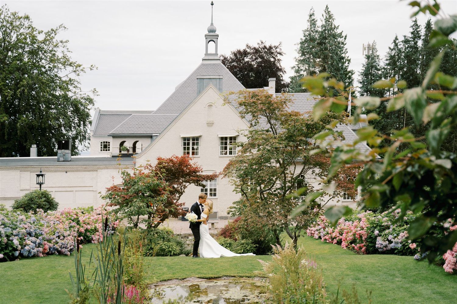 Bride and groom embrace in lush garden with hydrangeas before a white chateau.
