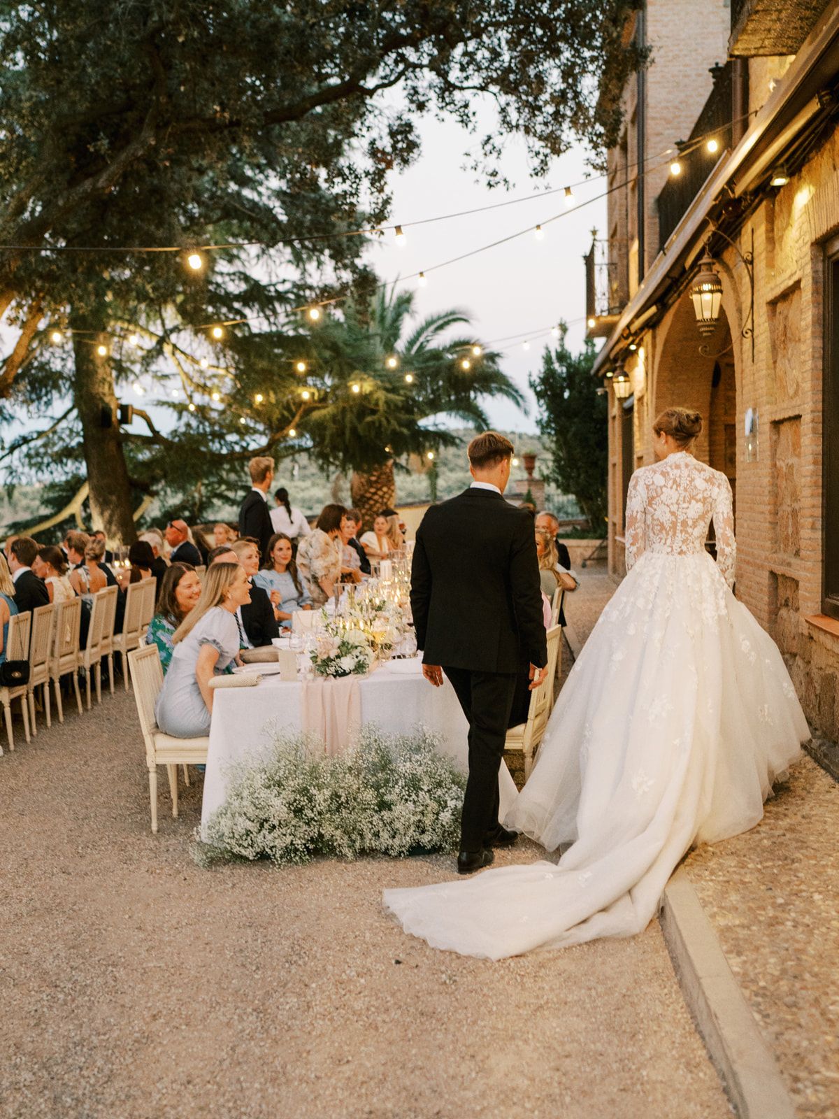 Bride and groom arrive at elegant outdoor wedding reception with string lights and long dining tables.